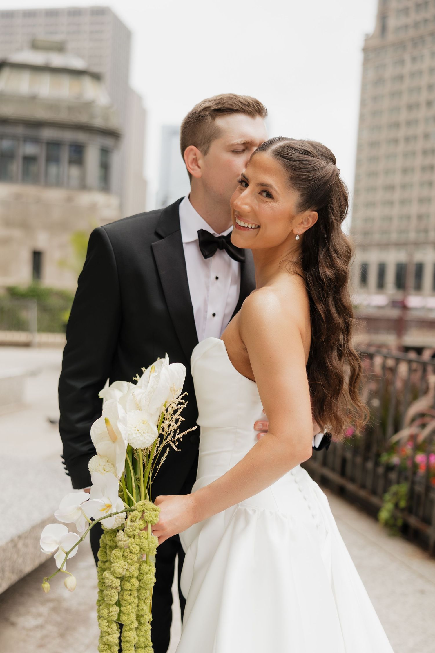 Groom in black tuxedo and bow tie kisses smiling bride in strapless white gown holding an orchid and green bouquet in a city setting.