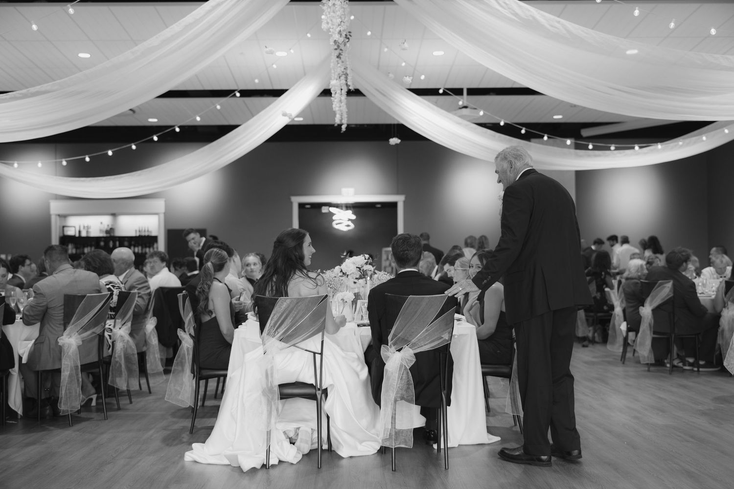 Black and white wedding reception photo showing bride and groom seated at head table with guests mingling in elegant draped venue.