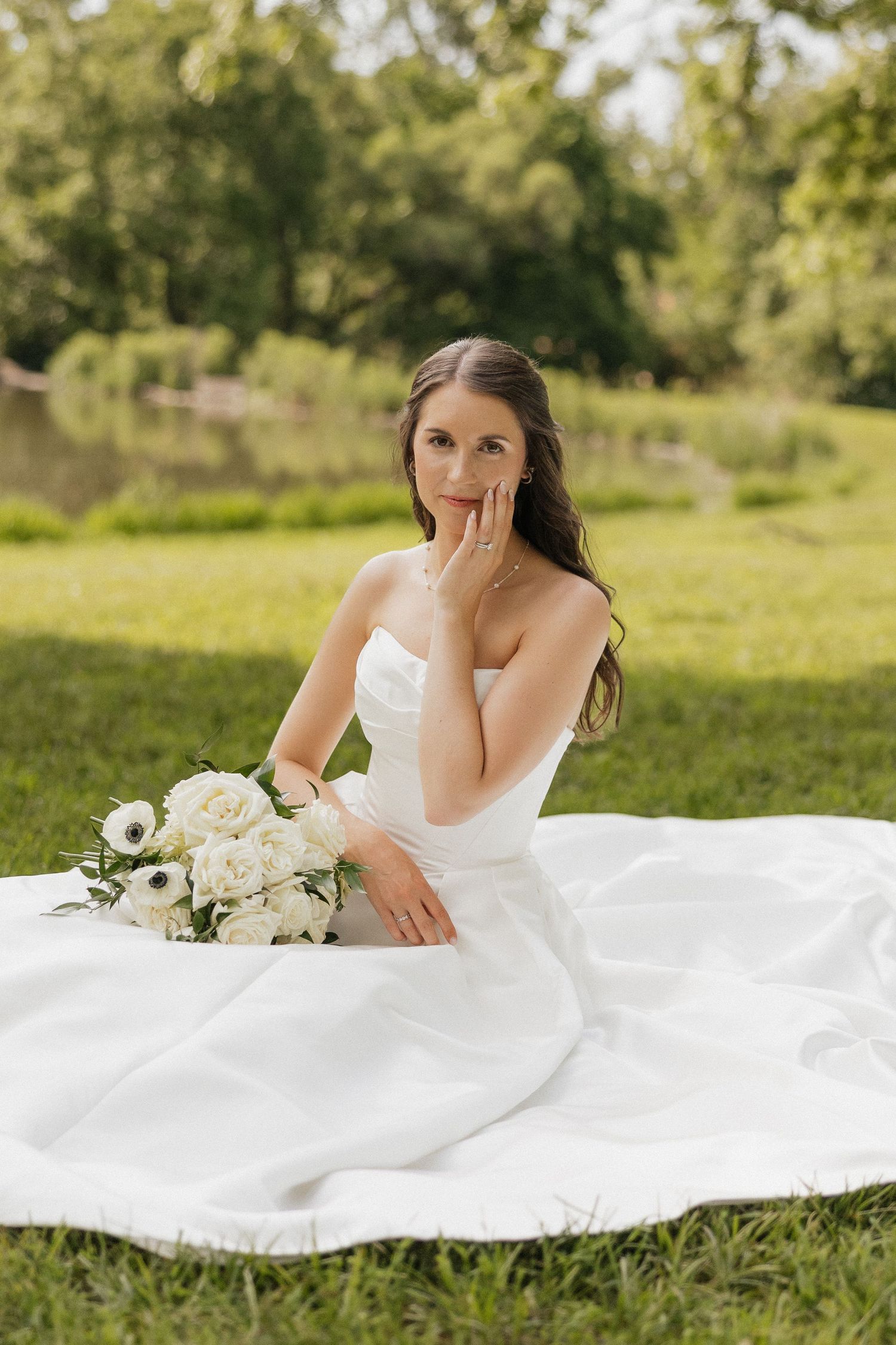 Bride in strapless white gown sits on grass holding white rose bouquet, posing elegantly in a lush green park setting.