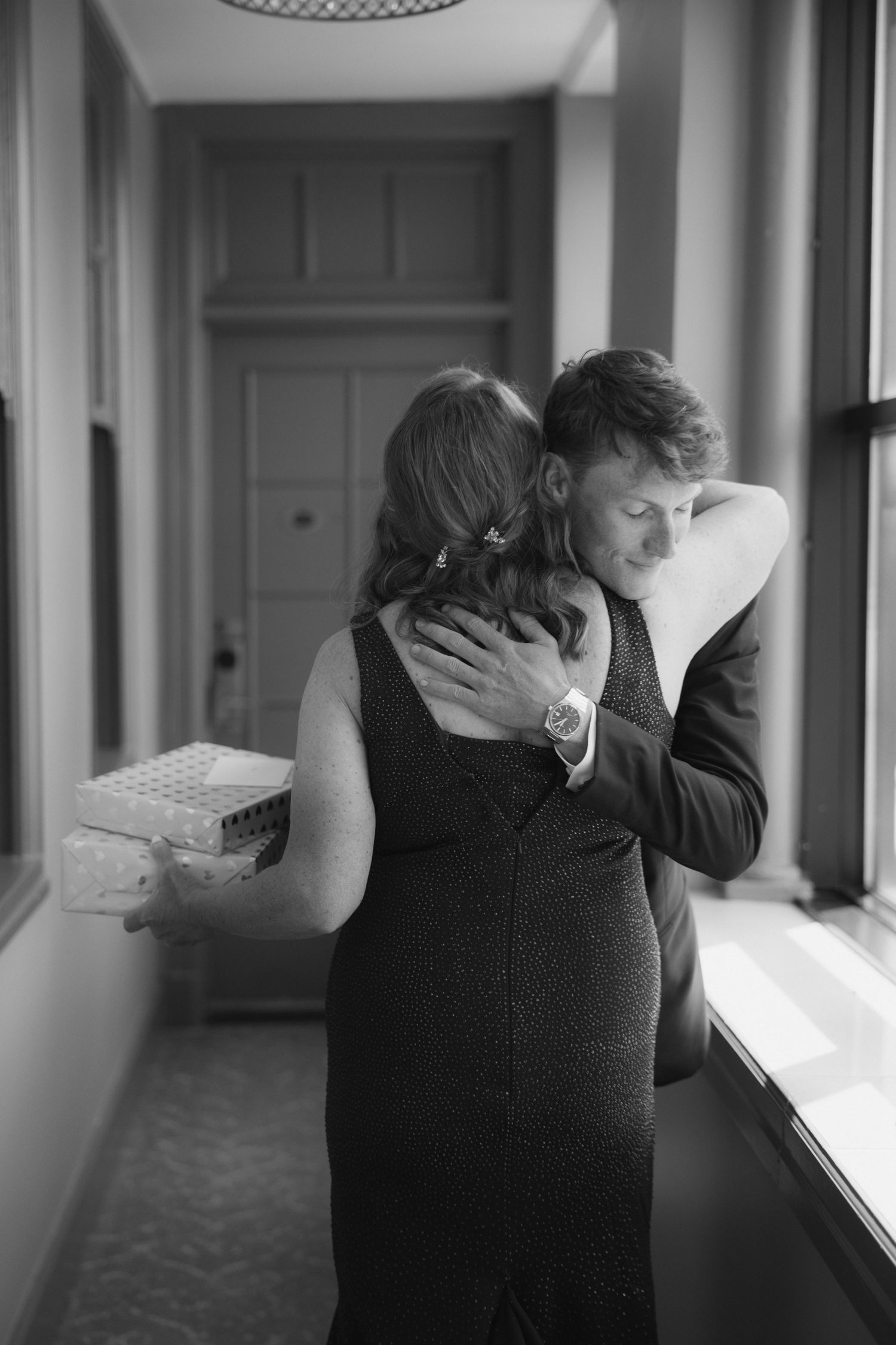 Black and white photo of a couple embracing tenderly in a hallway, woman holding a gift box.