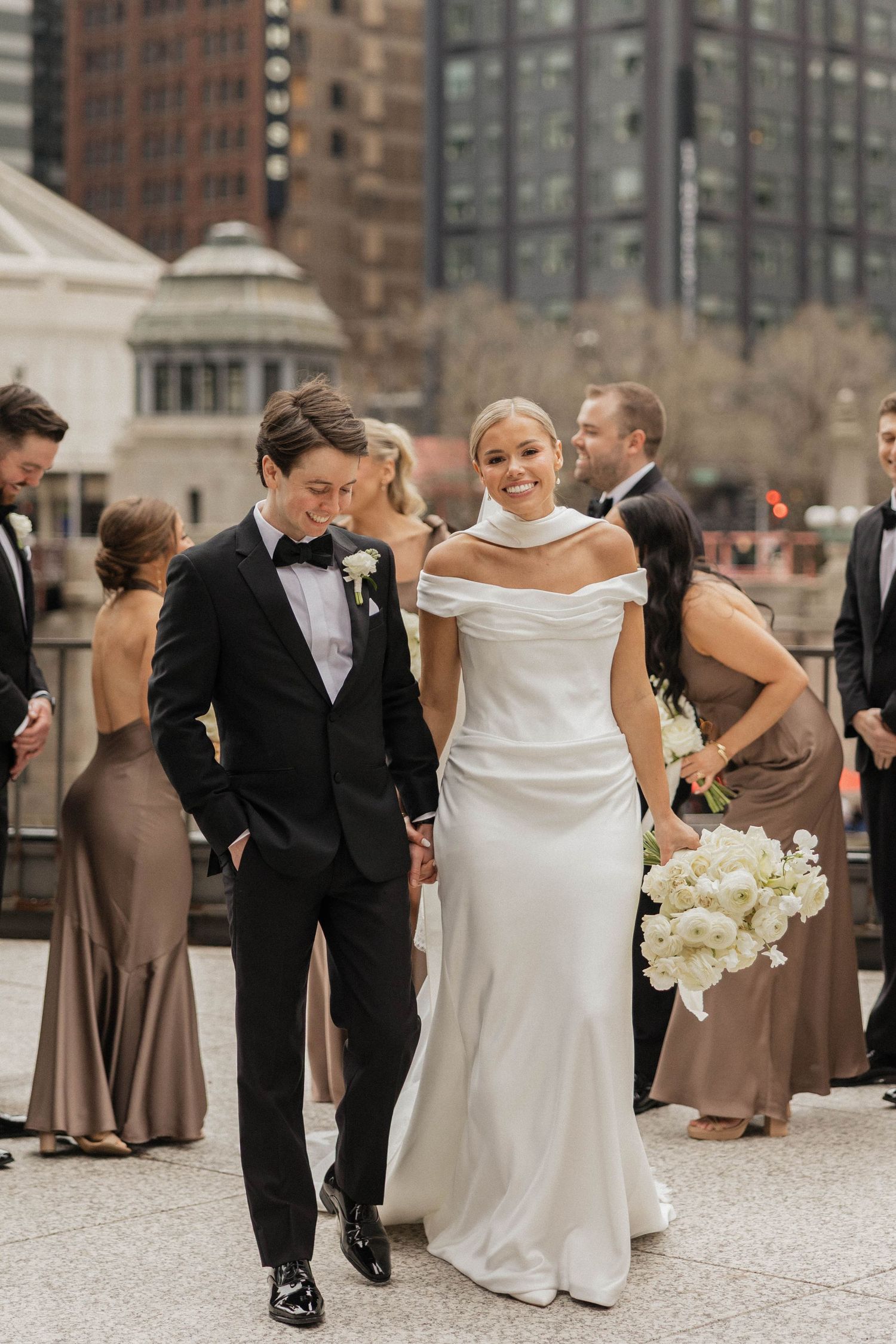 Smiling bride in off-shoulder gown walks with tuxedo groom at Chicago outdoor venue.