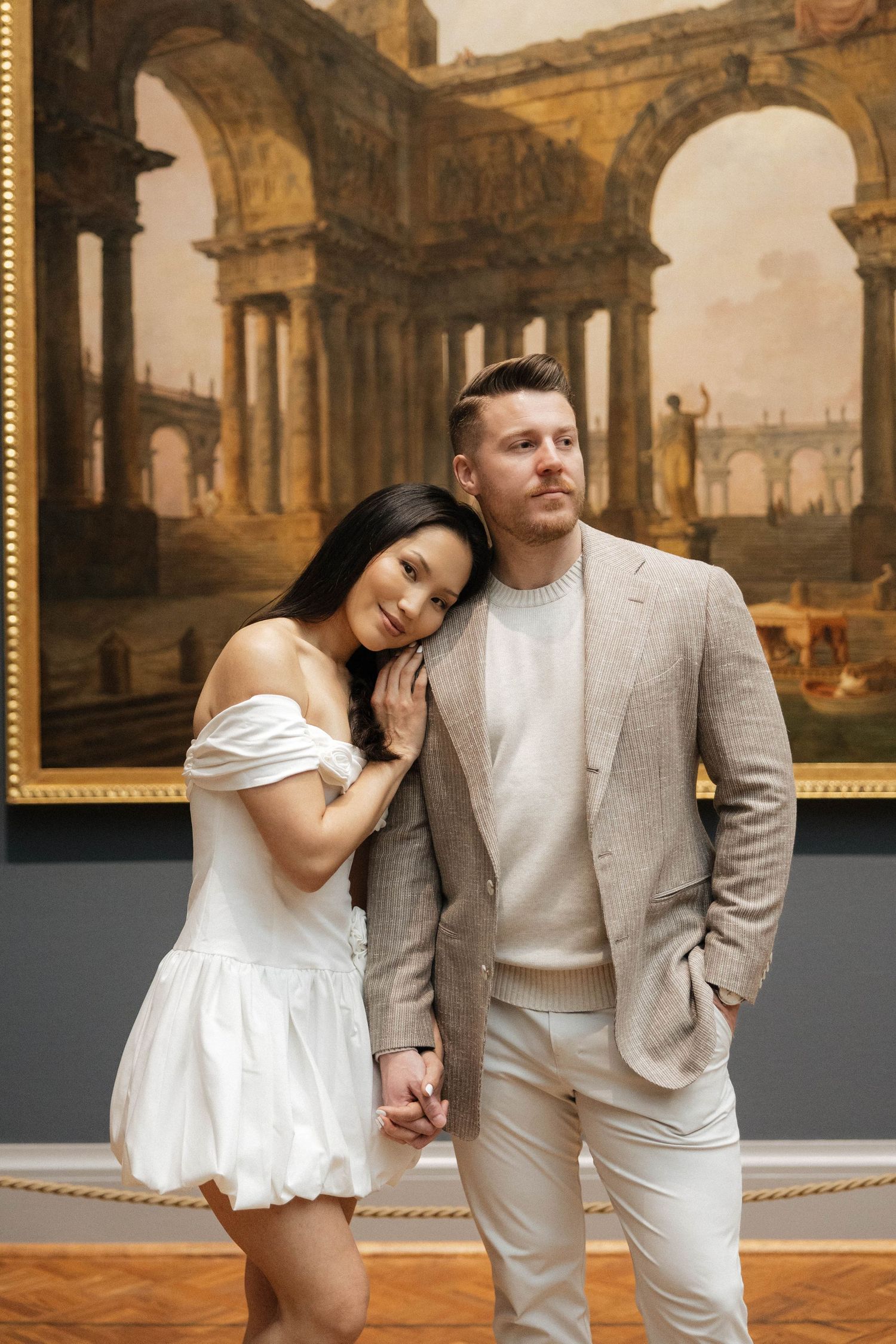 Elegant couple posing in an art museum in front of a classical painting, wearing neutral tones and white dress.