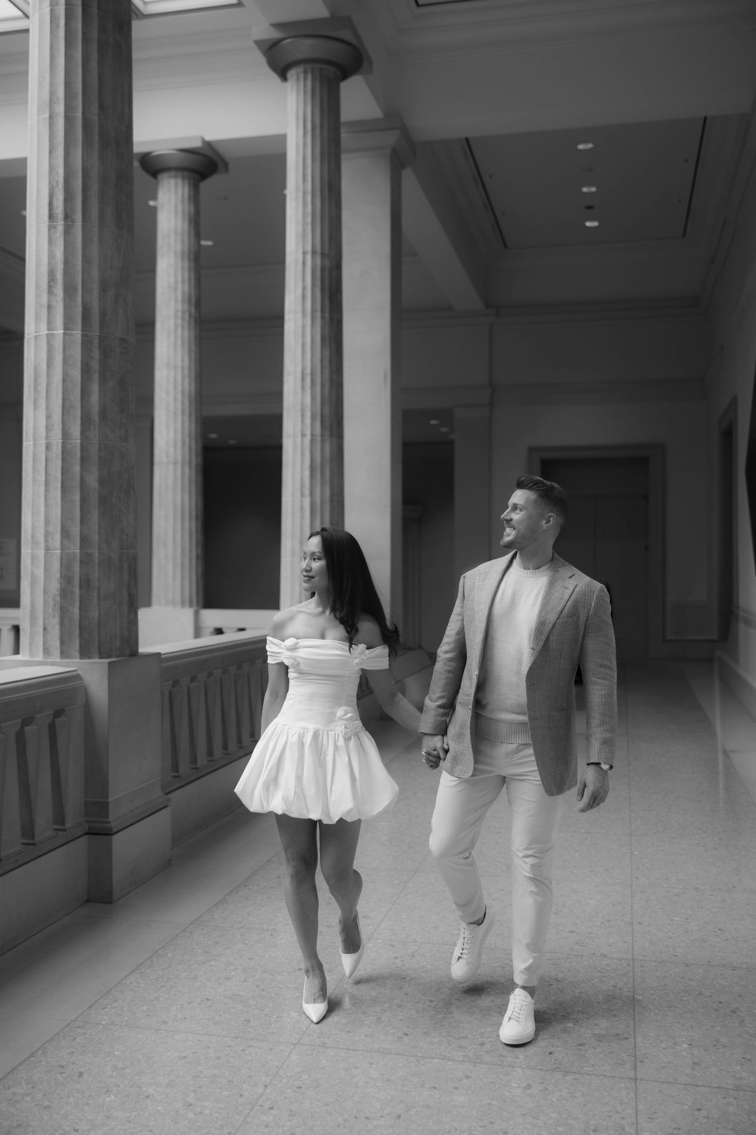 Black and white photo of an elegant couple holding hands, walking through a classical columned hallway.