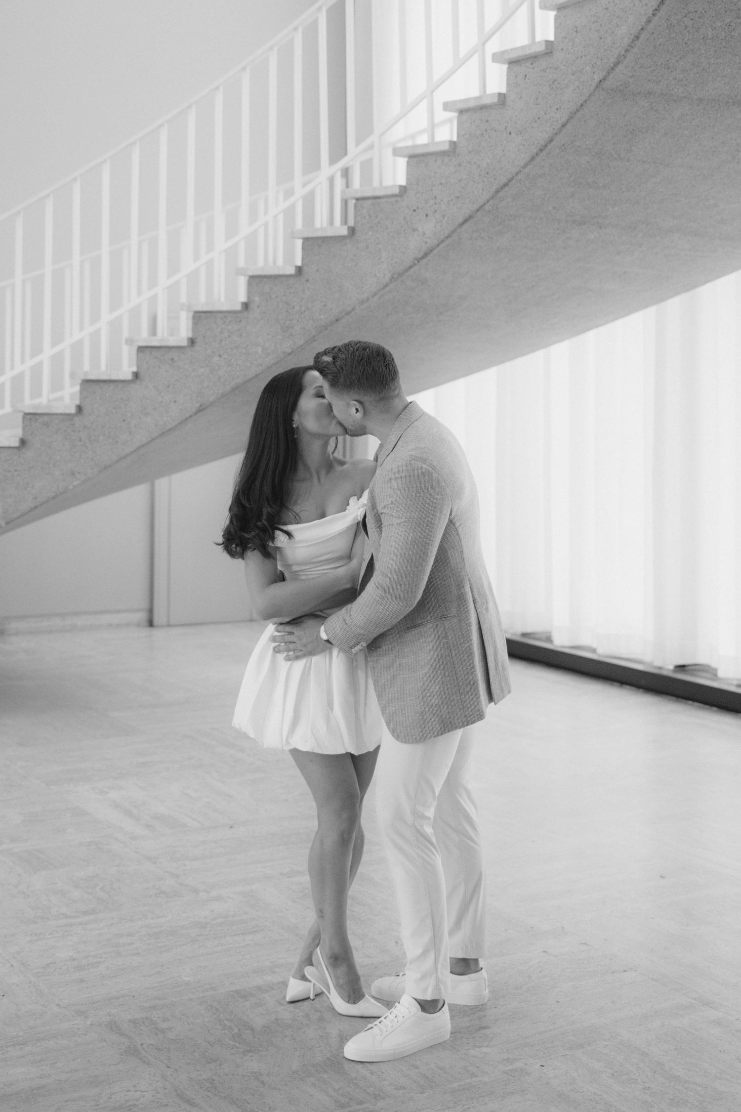 Black and white photo of a couple sharing a romantic kiss in an elegant interior with a curved staircase.