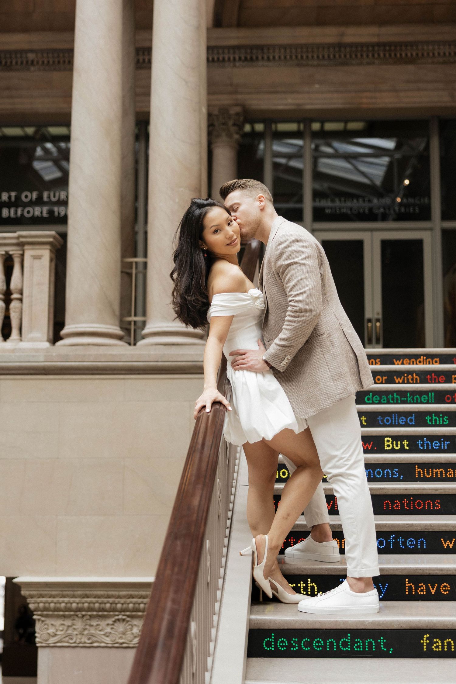 Couple sharing a romantic kiss on colorful text-adorned steps of a grand building with columns.