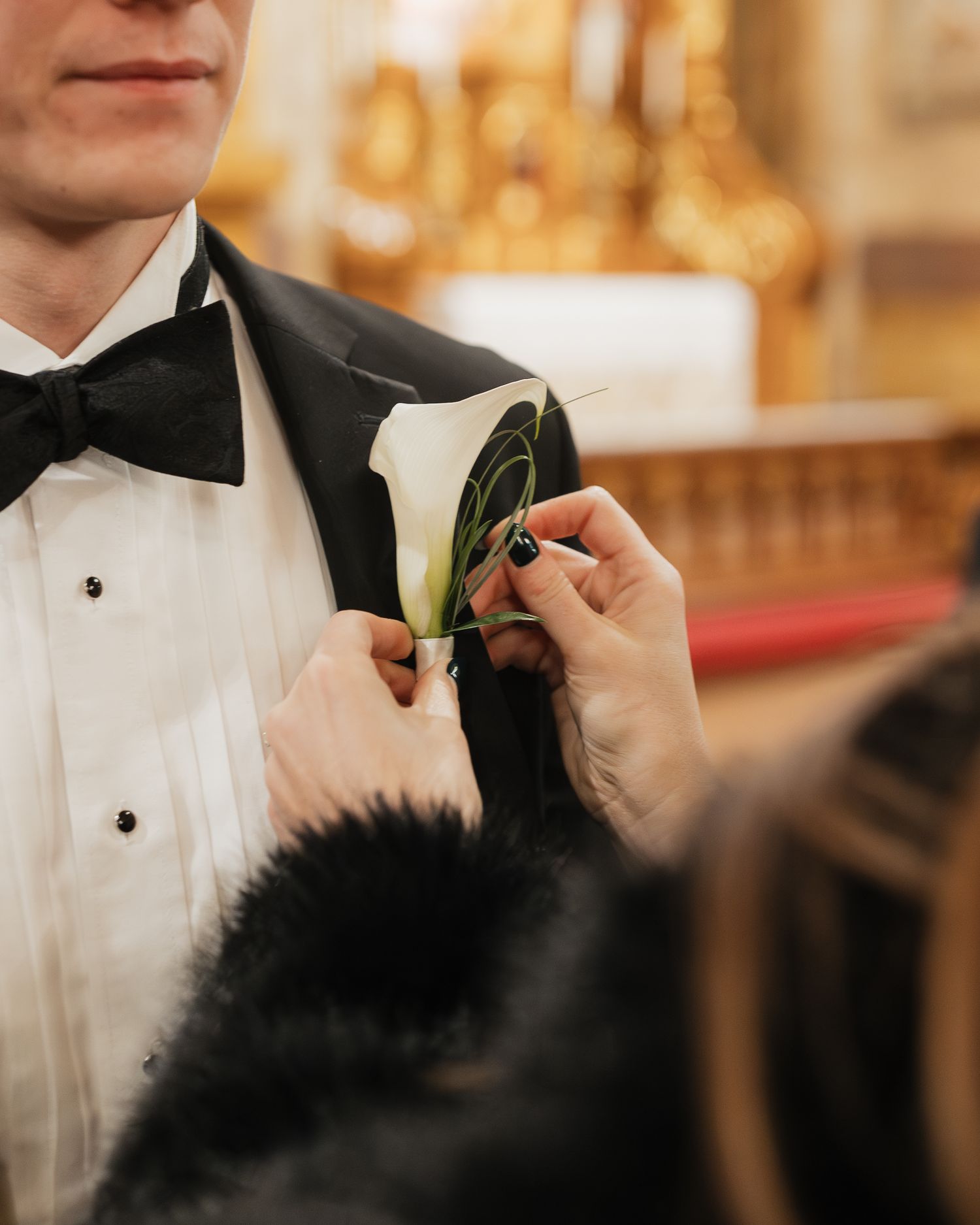 Close-up of hands pinning a white boutonniere onto a groom's black tuxedo lapel.