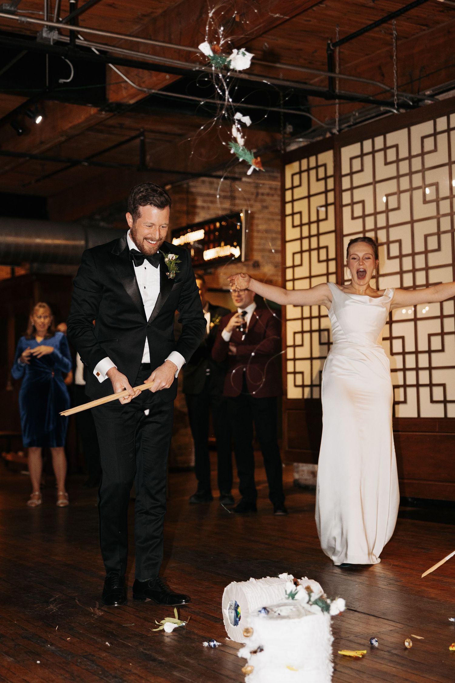 Wedding couple participates in cake smashing tradition at reception with geometric wall decor and exposed ceiling.