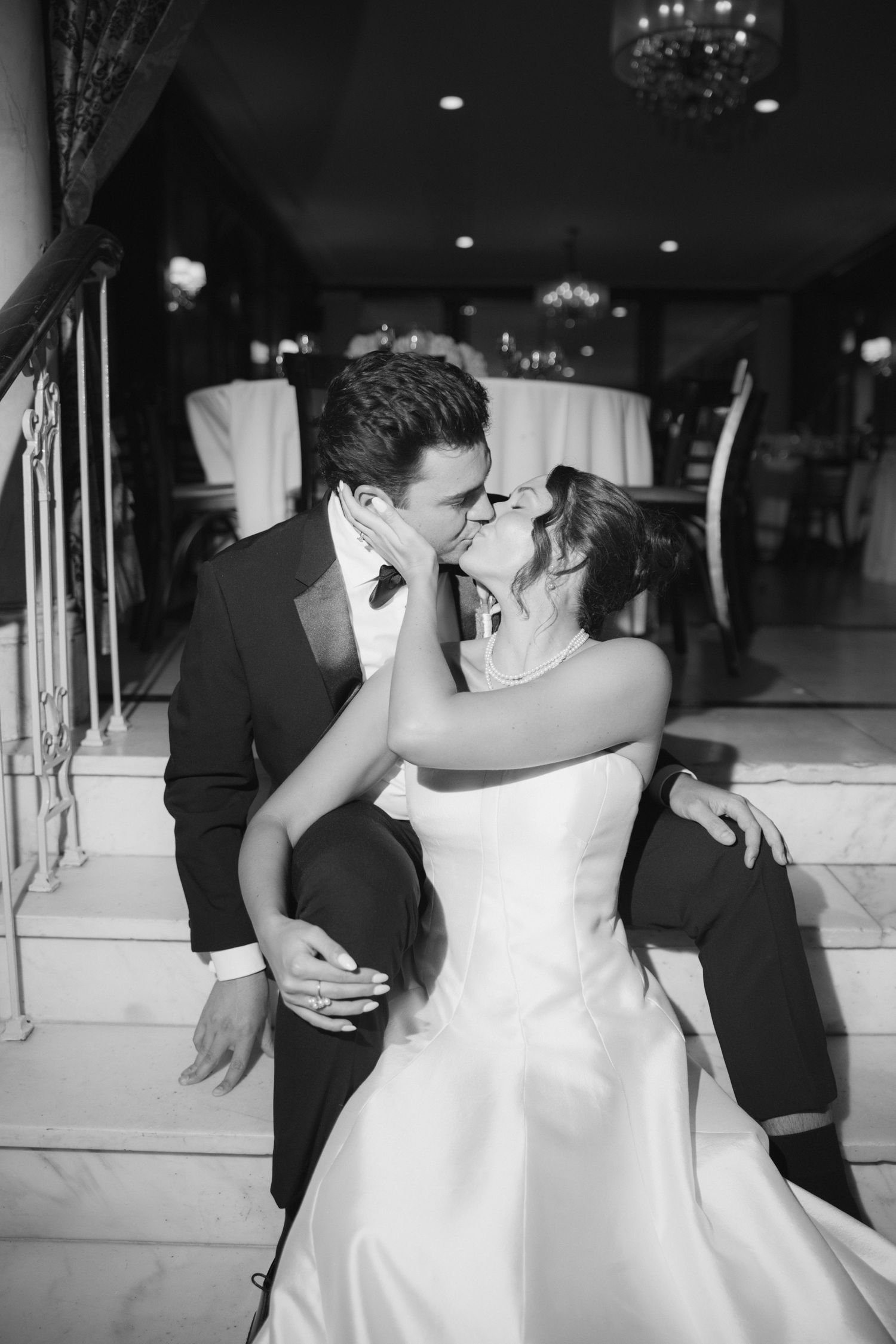 Black and white photo of a bride and groom sharing a romantic kiss on steps at their elegant wedding reception.