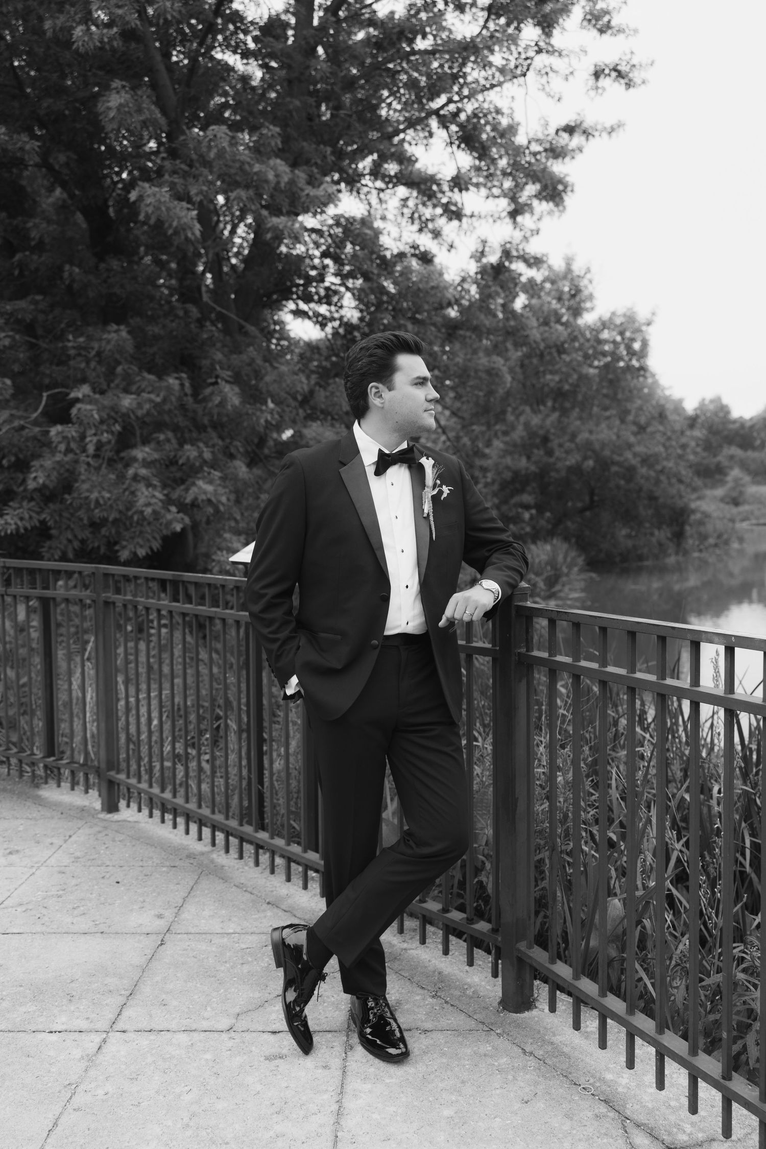 Black and white photo of a groom in a tuxedo leaning on a railing outdoors with trees and water in the background.