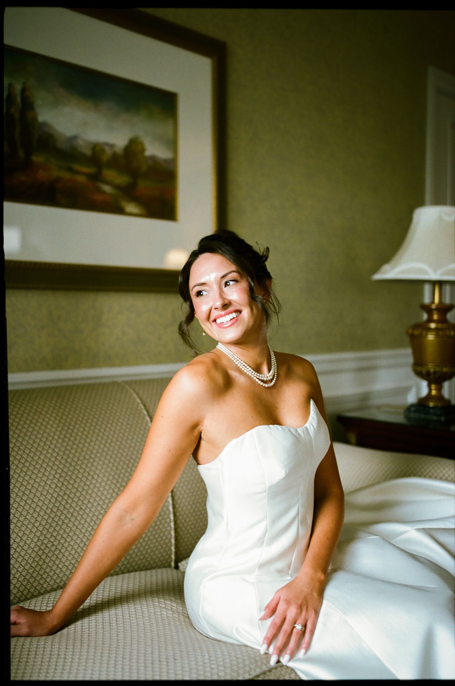Smiling bride in strapless white gown and pearl necklace sitting on a sofa in an elegant hotel room.