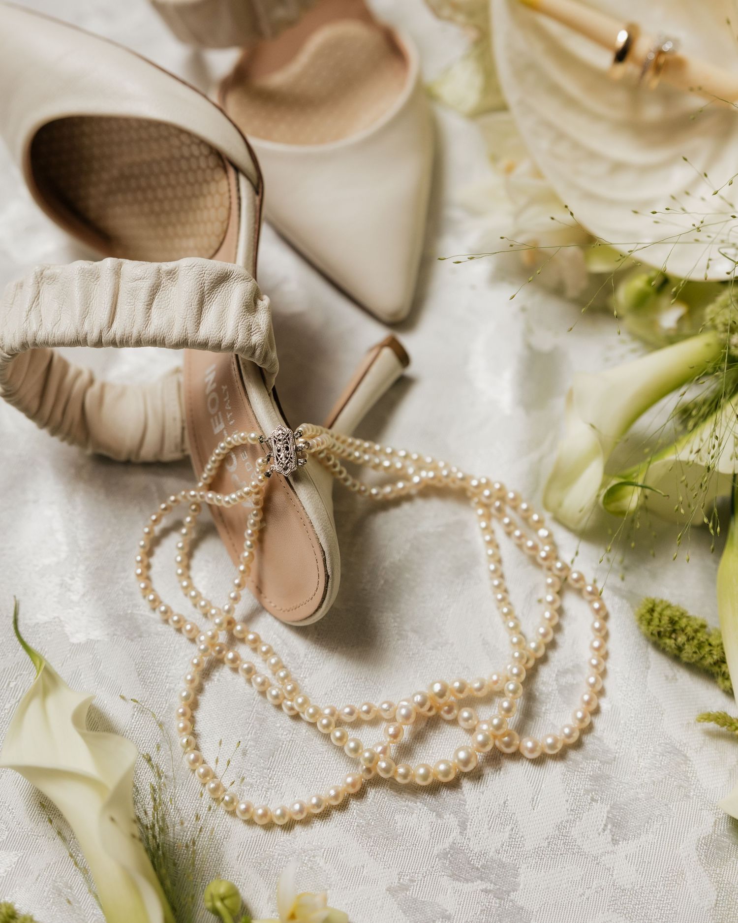 Elegant bridal flat lay featuring pearl necklace, ivory satin heels, and white calla lilies on lace fabric.