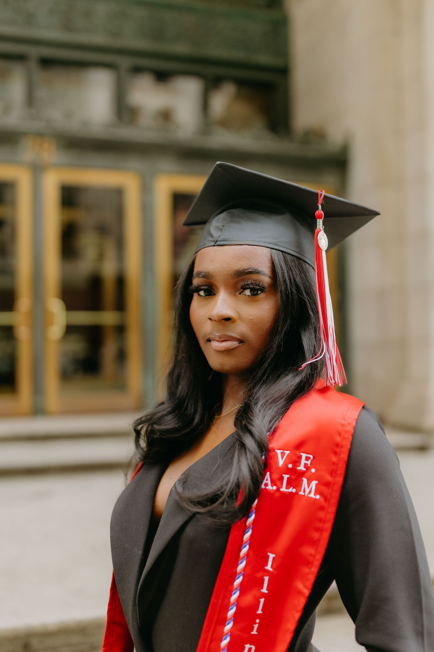 A graduate wearing a black cap and gown with a red graduation stole poses outdoors near a building entrance.