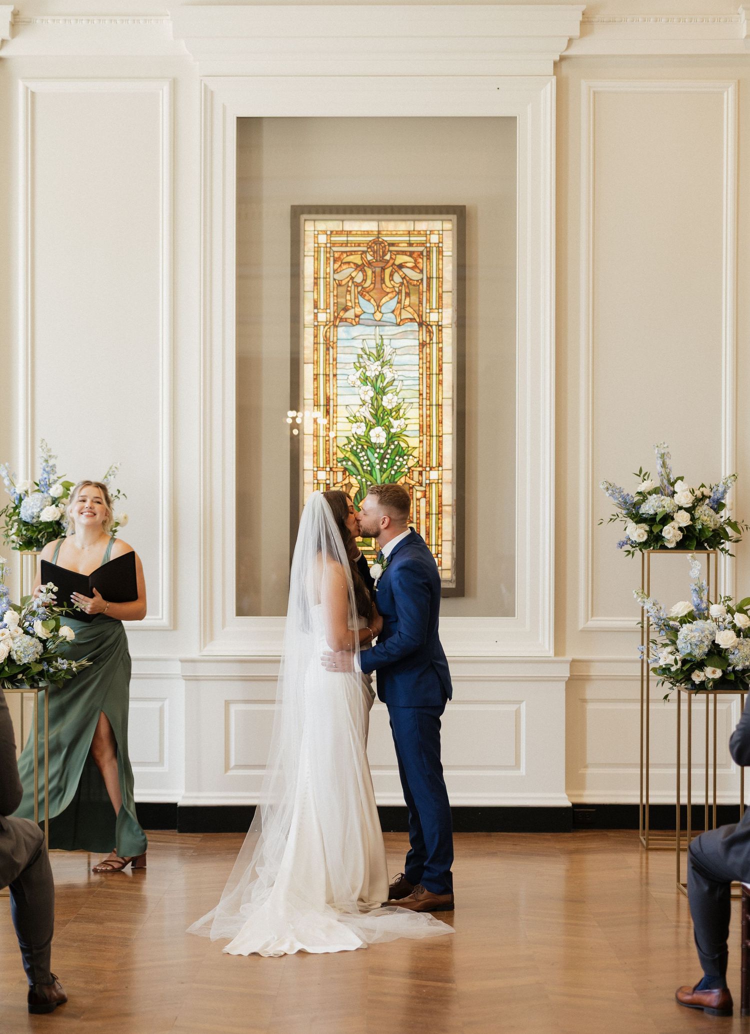 An elegant wedding ceremony takes place in a bright room with white walls and a stained glass window backdrop.