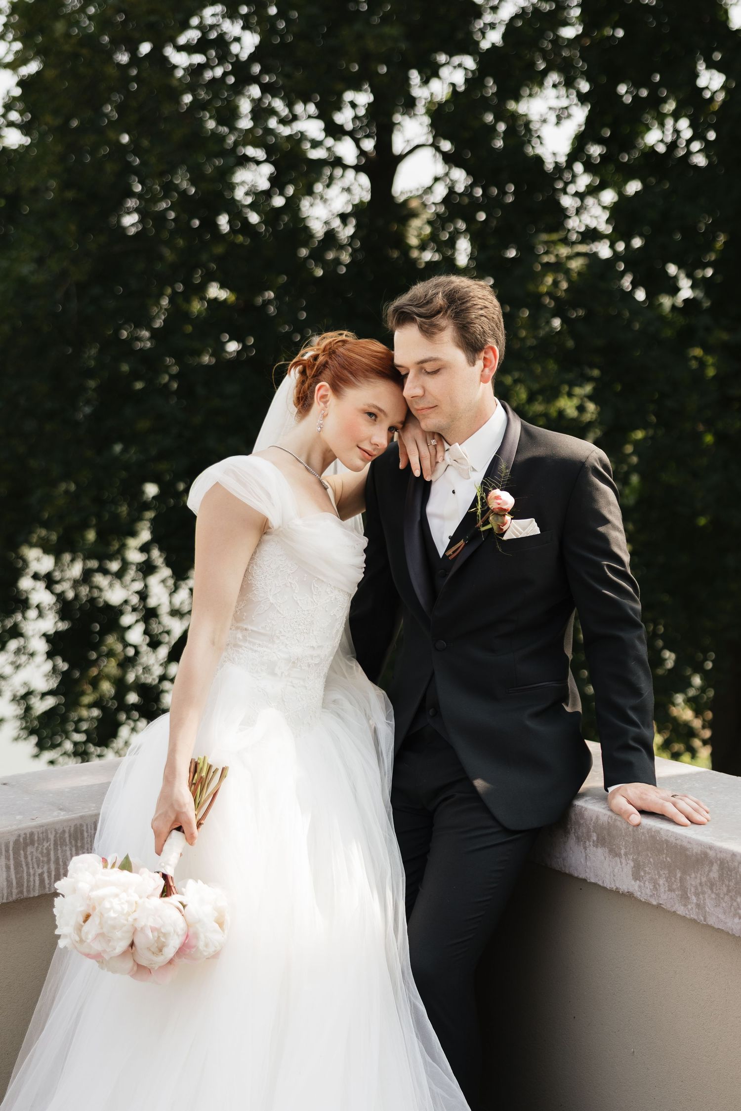 Bride and groom share a tender moment on a balcony, she in white lace gown holding peonies, he in dark suit.