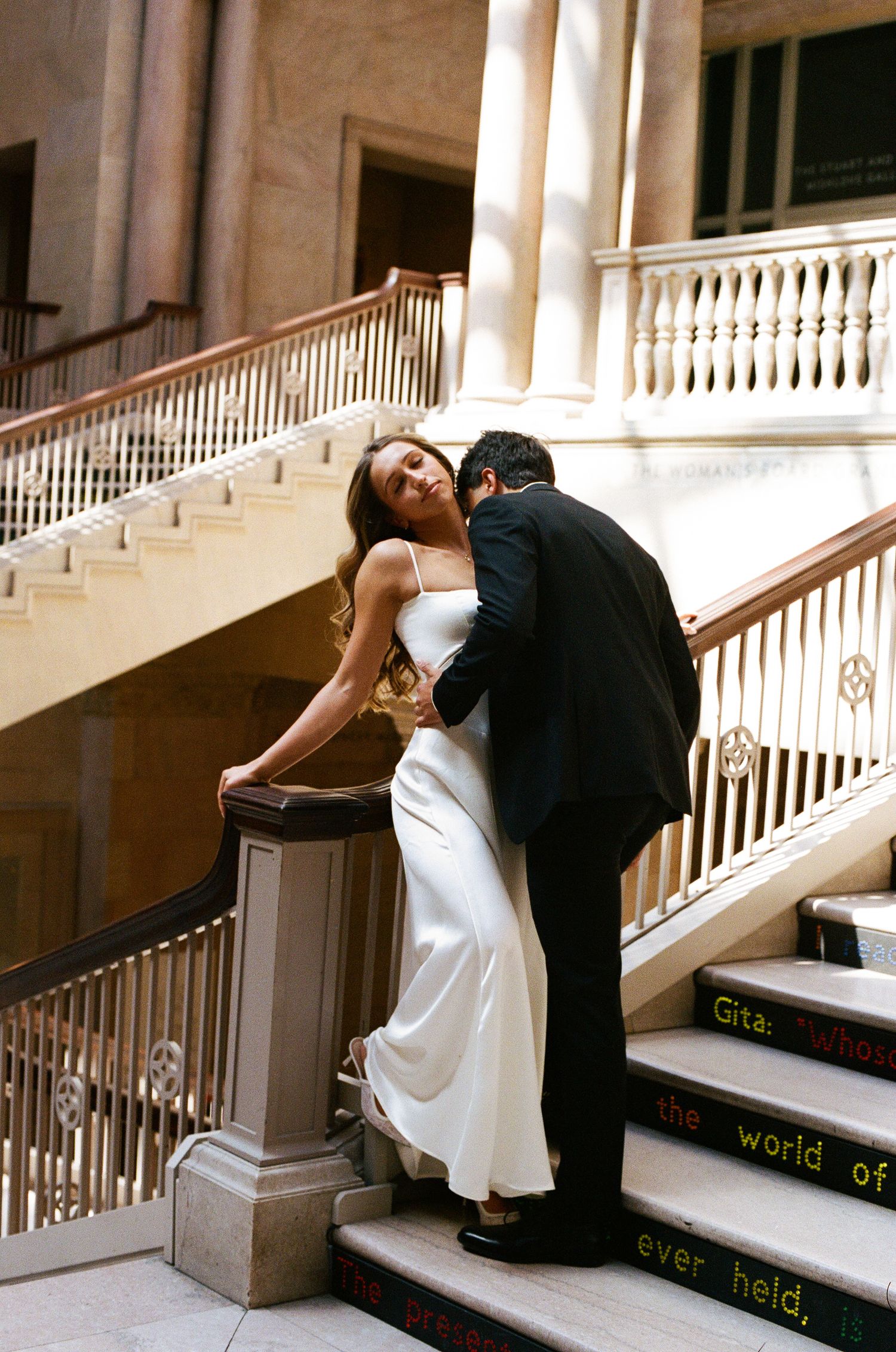 Bride in white gown and groom in black suit share a romantic kiss on grand staircase of elegant venue.