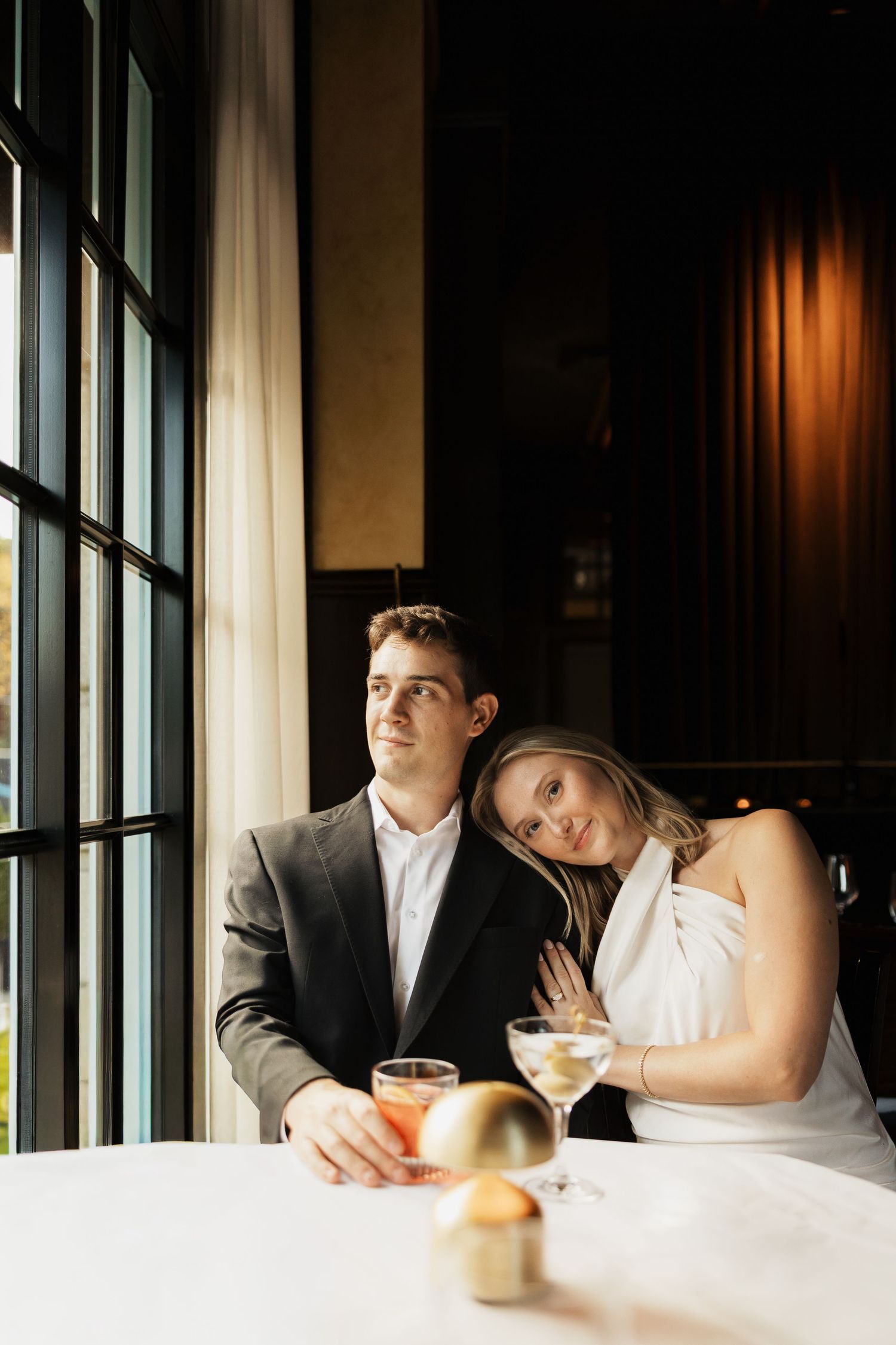 Smiling woman in a white dress rests her head on a man's shoulder at a elegant restaurant table with cocktails by a tall window.