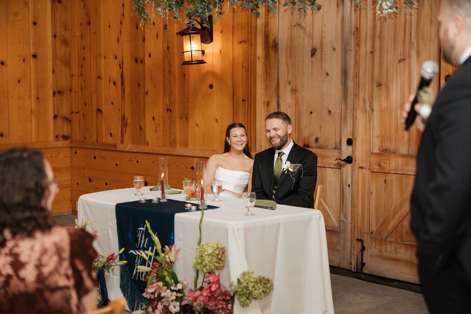 Smiling bride and groom sit at a white-draped head table with flowers in a rustic wooden venue while a guest gives a toast.