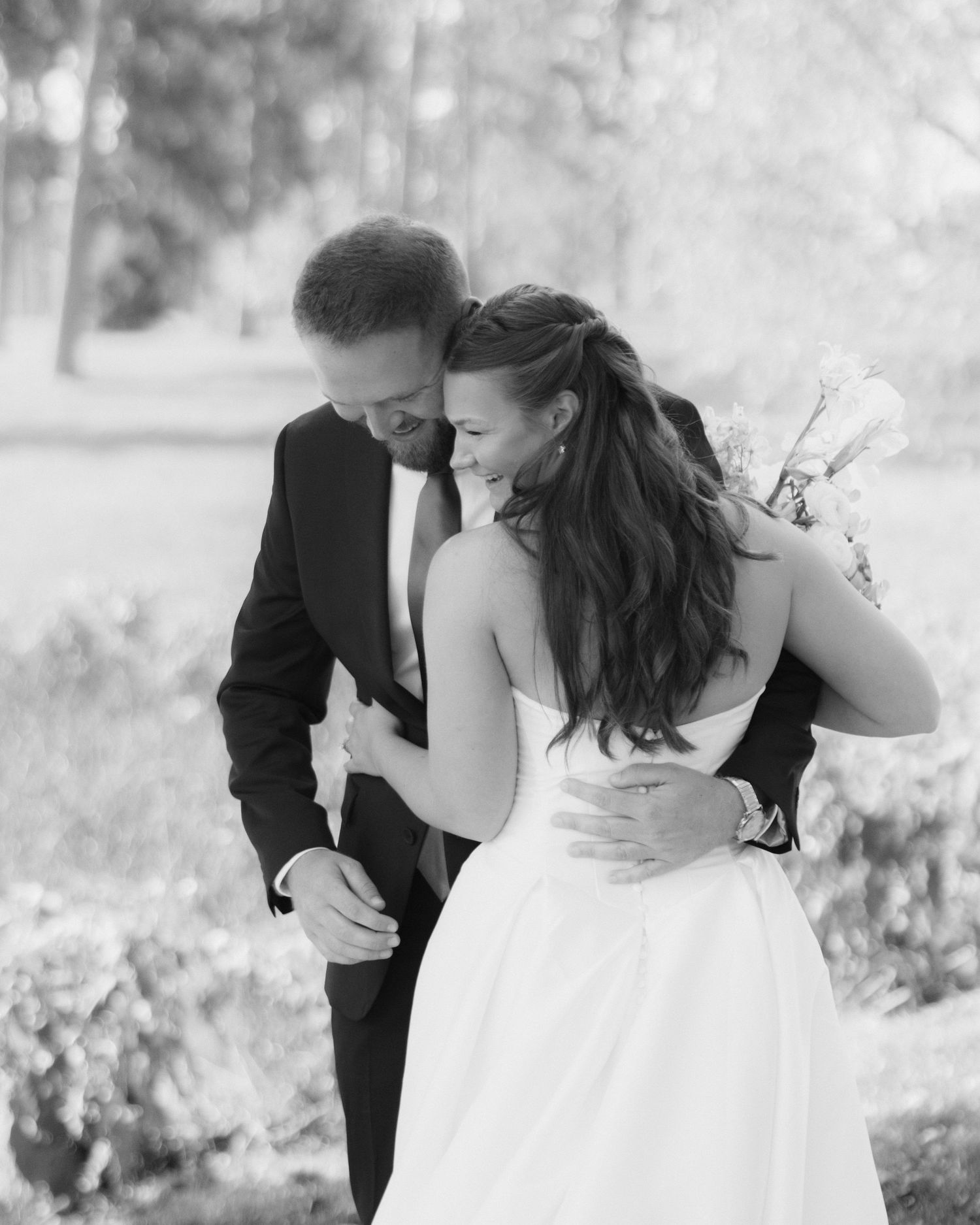 Black and white photo of a bride in a strapless gown embracing a groom in a suit outdoors, holding a bouquet among soft green trees.