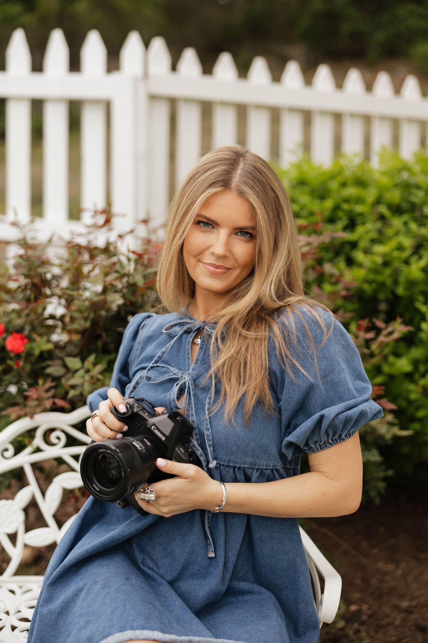 Blonde woman in blue denim dress holding a camera, sitting on white bench in garden with white picket fence.