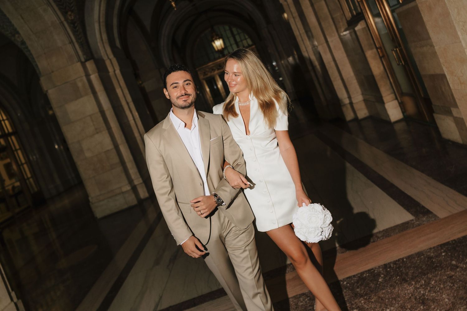 Couple in wedding attire poses at night, man in tan suit, woman in white mini dress holding floral bouquet.