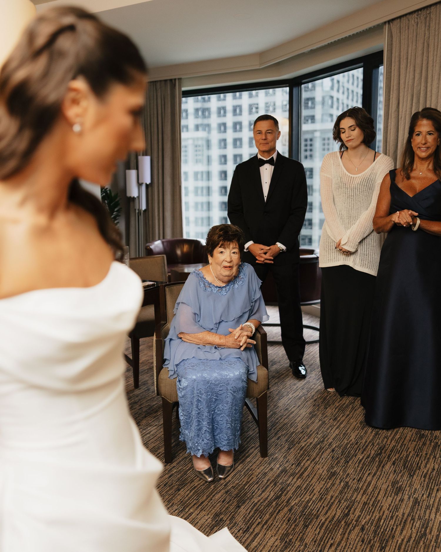 Bride in white gown stands before emotional family members, including seated elderly woman, in elegant hotel room.