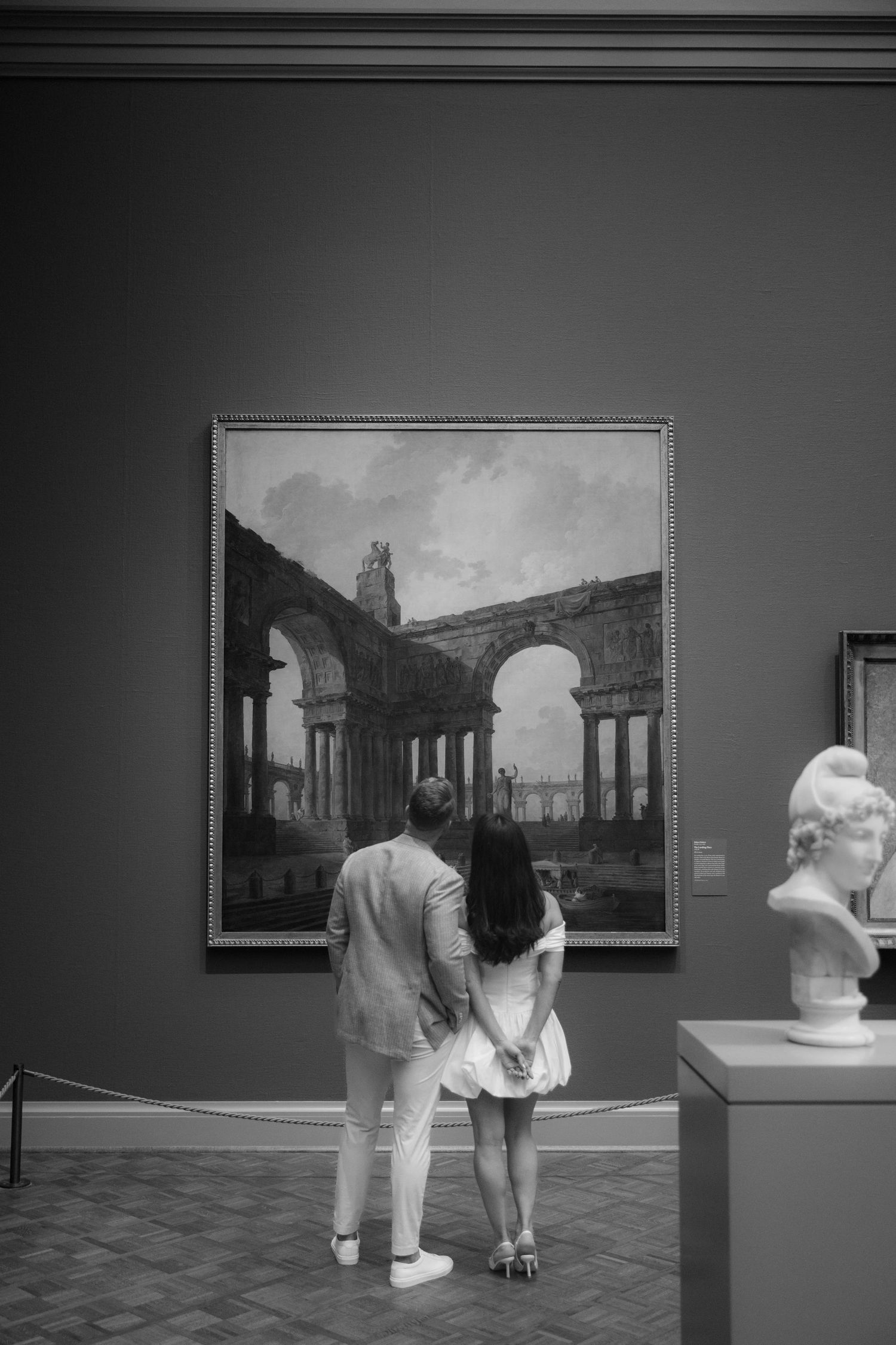 Couple viewing a large architectural painting in a museum gallery with a marble bust sculpture nearby.