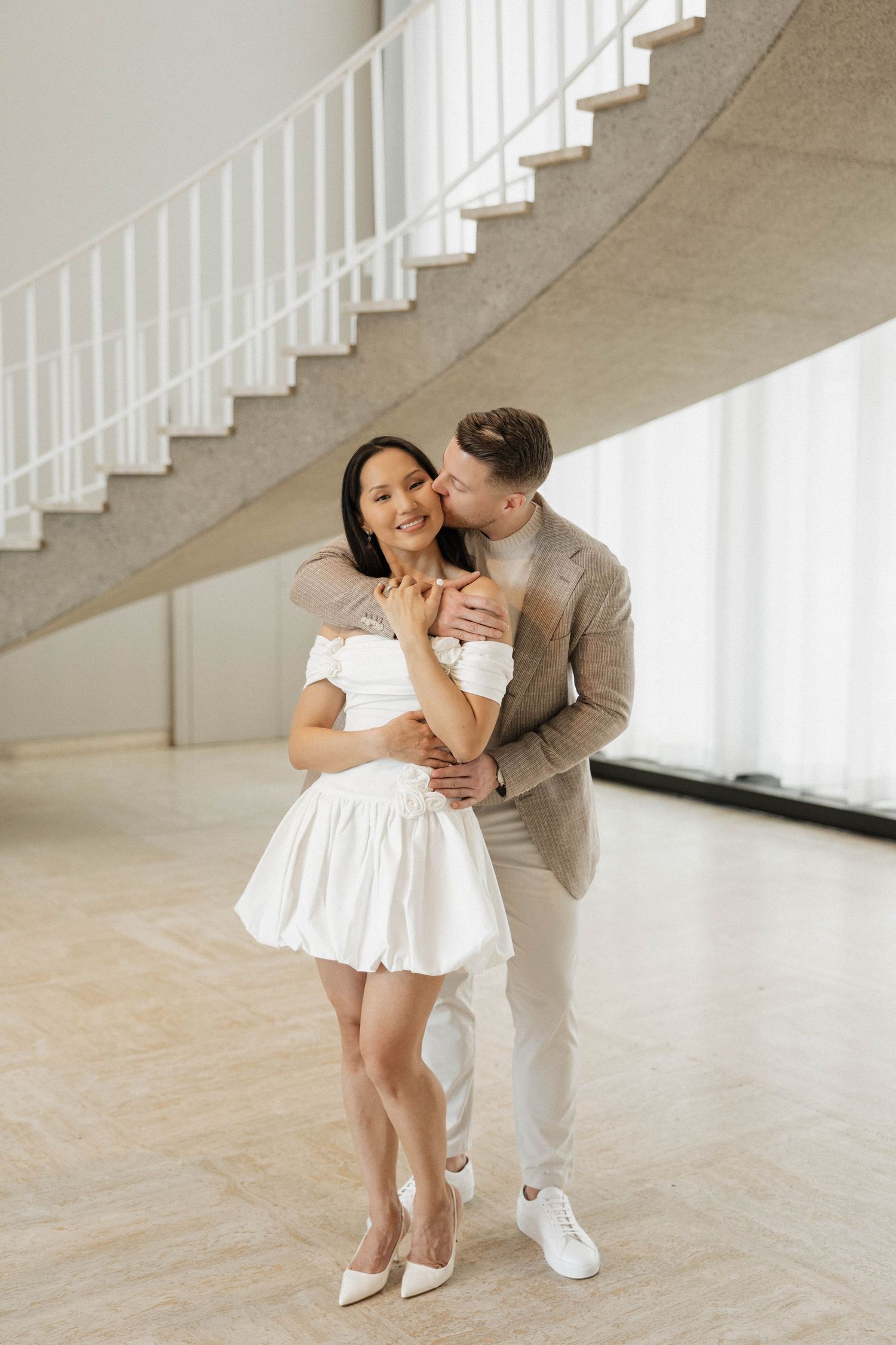 Elegant couple posing in modern venue, woman in white mini dress, man in beige suit embracing her near spiral staircase.