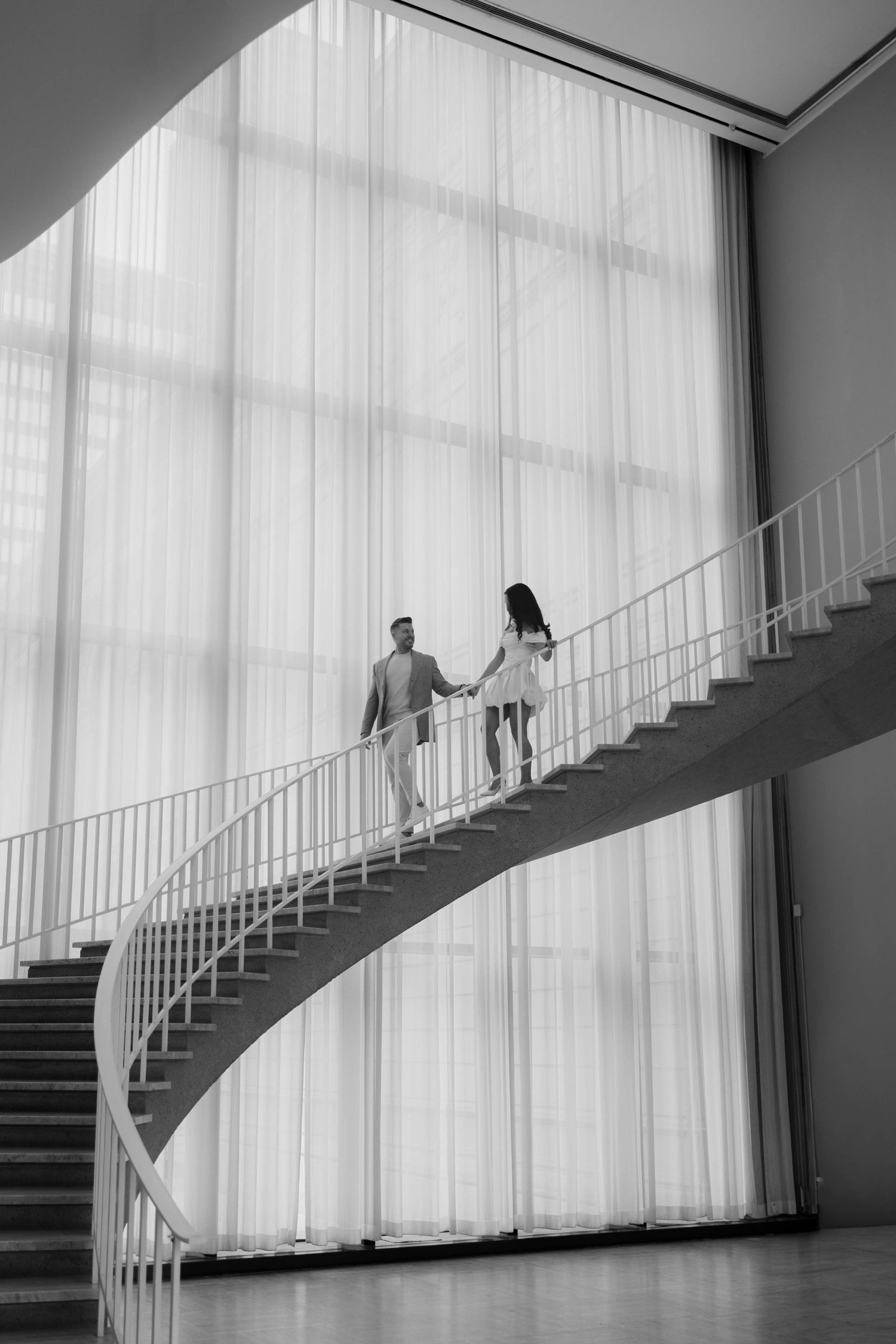 Black and white photo of a couple holding hands on an elegant spiral staircase in a modern building.
