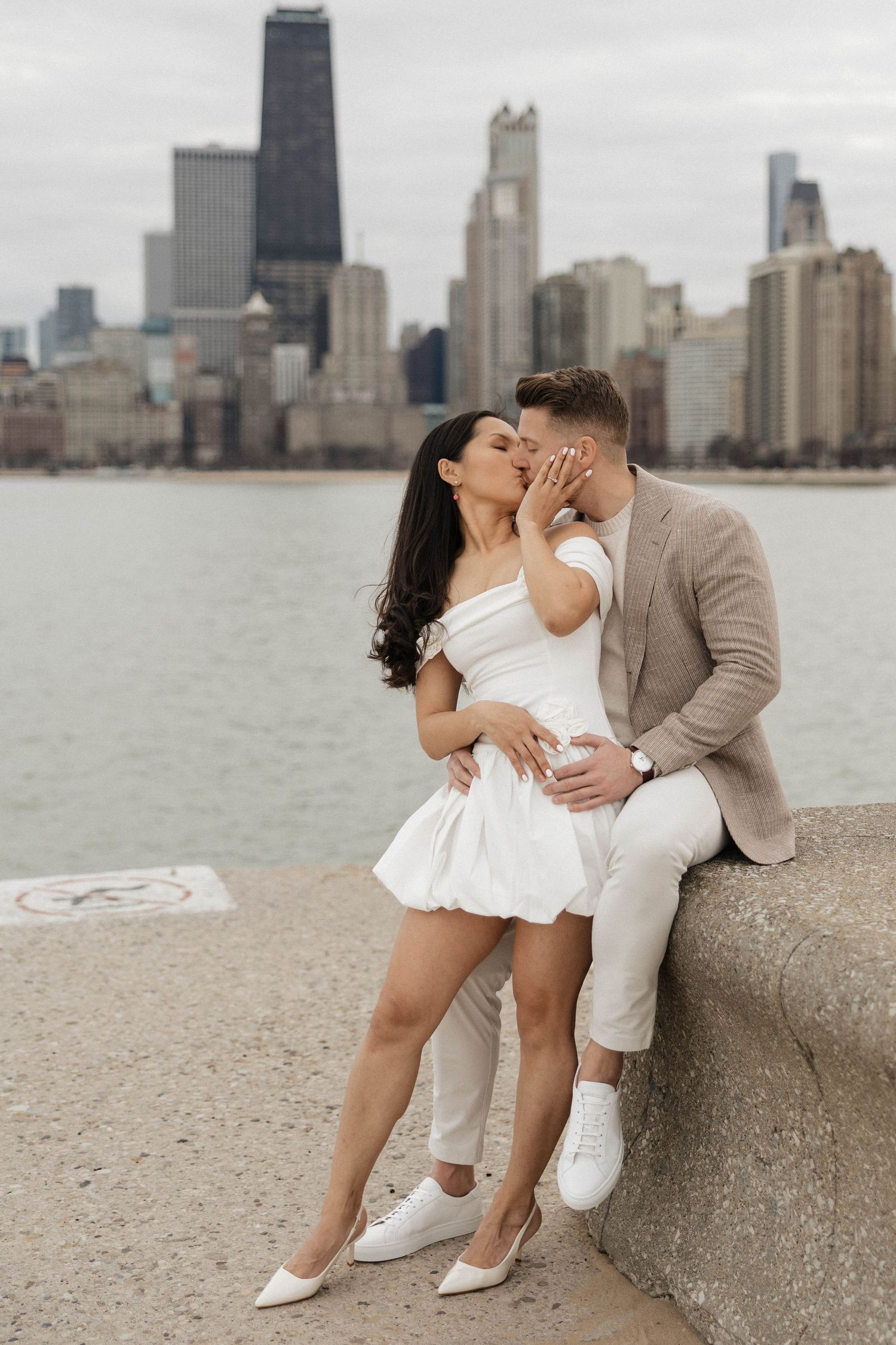Couple sharing a romantic kiss by the Chicago lakefront, woman in white mini dress, man in beige blazer, city skyline behind.
