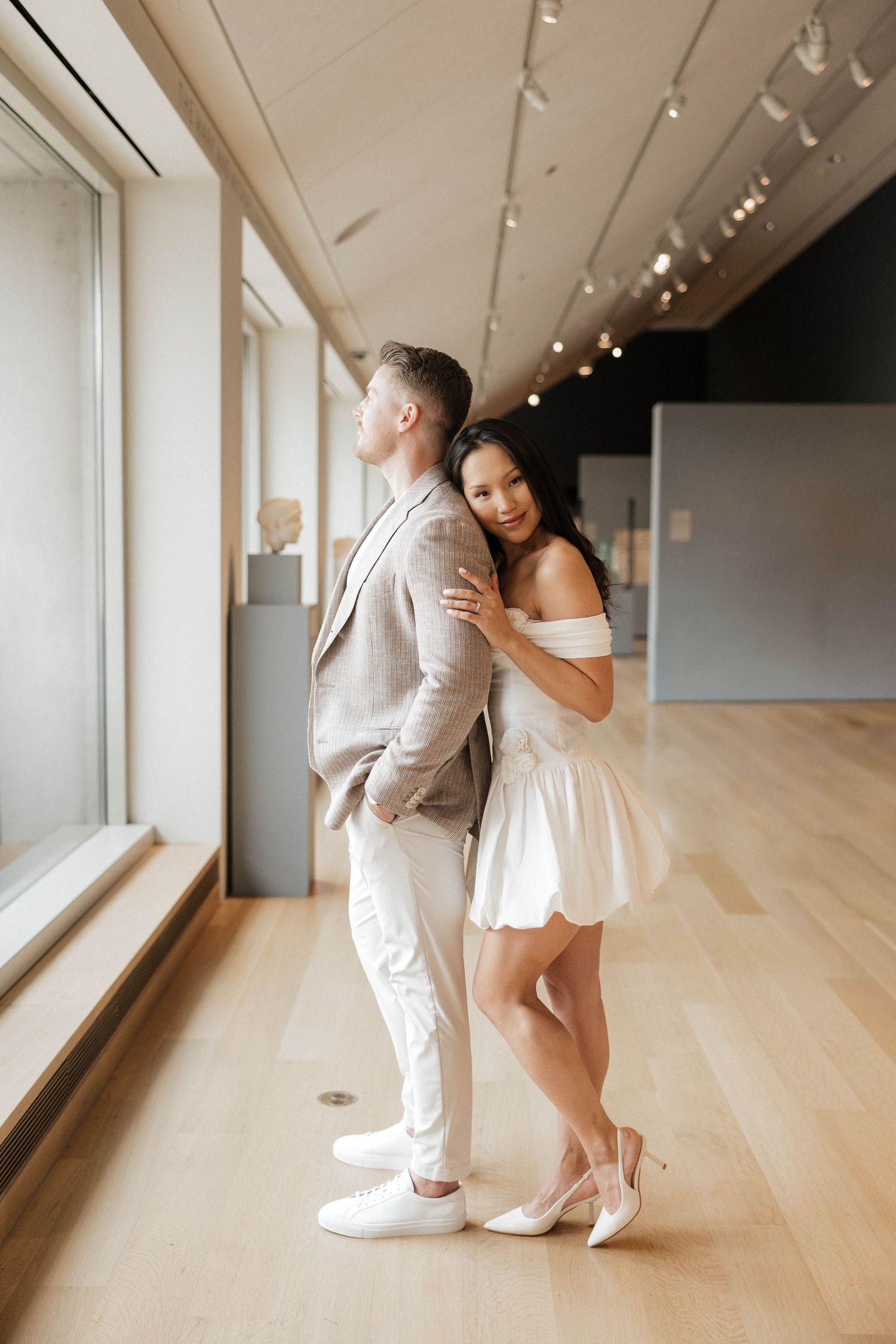 Couple posing in art museum gallery, woman in white mini dress embracing man in gray suit on hardwood floors.