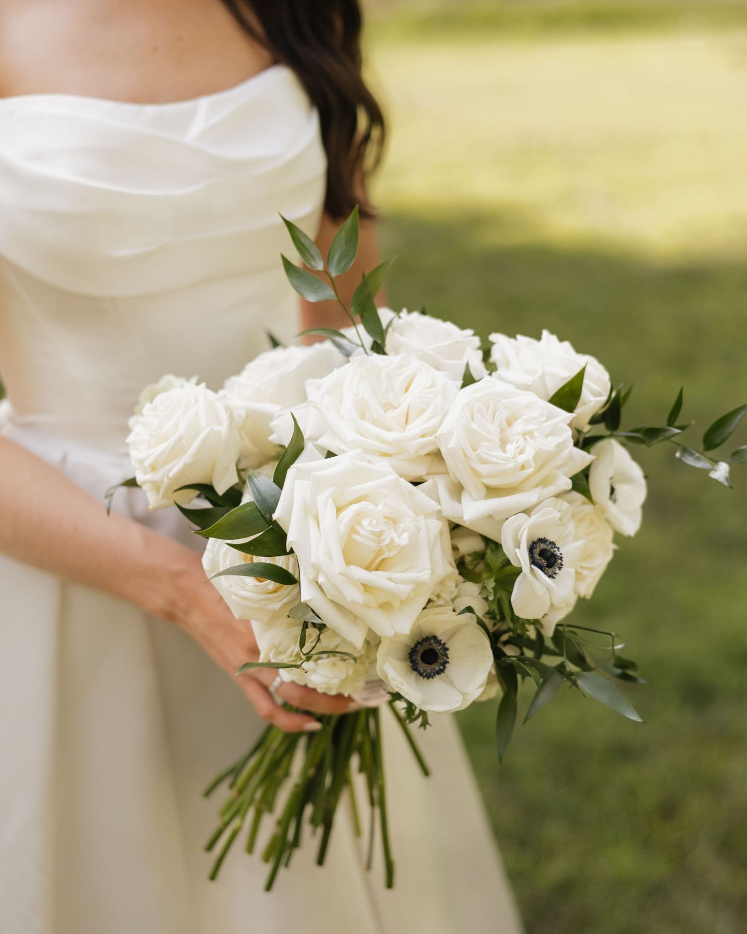 Bride holding white rose and anemone wedding bouquet with greenery accents in an elegant strapless gown.