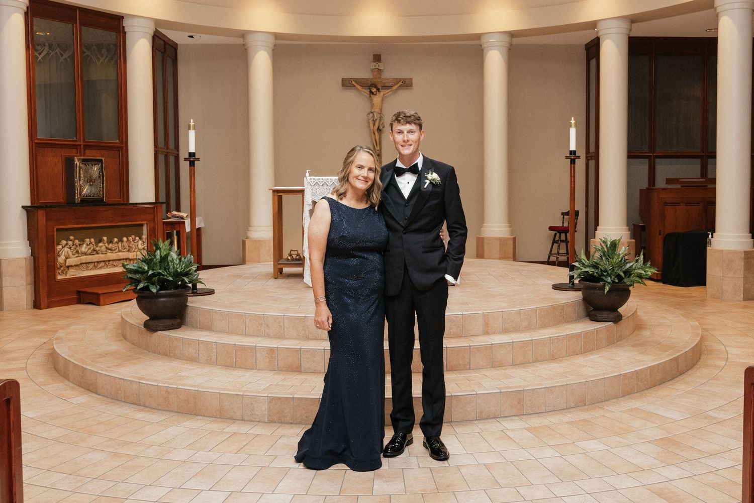 A woman in a navy gown and young man in a tuxedo pose together in an elegant church chapel with a crucifix.