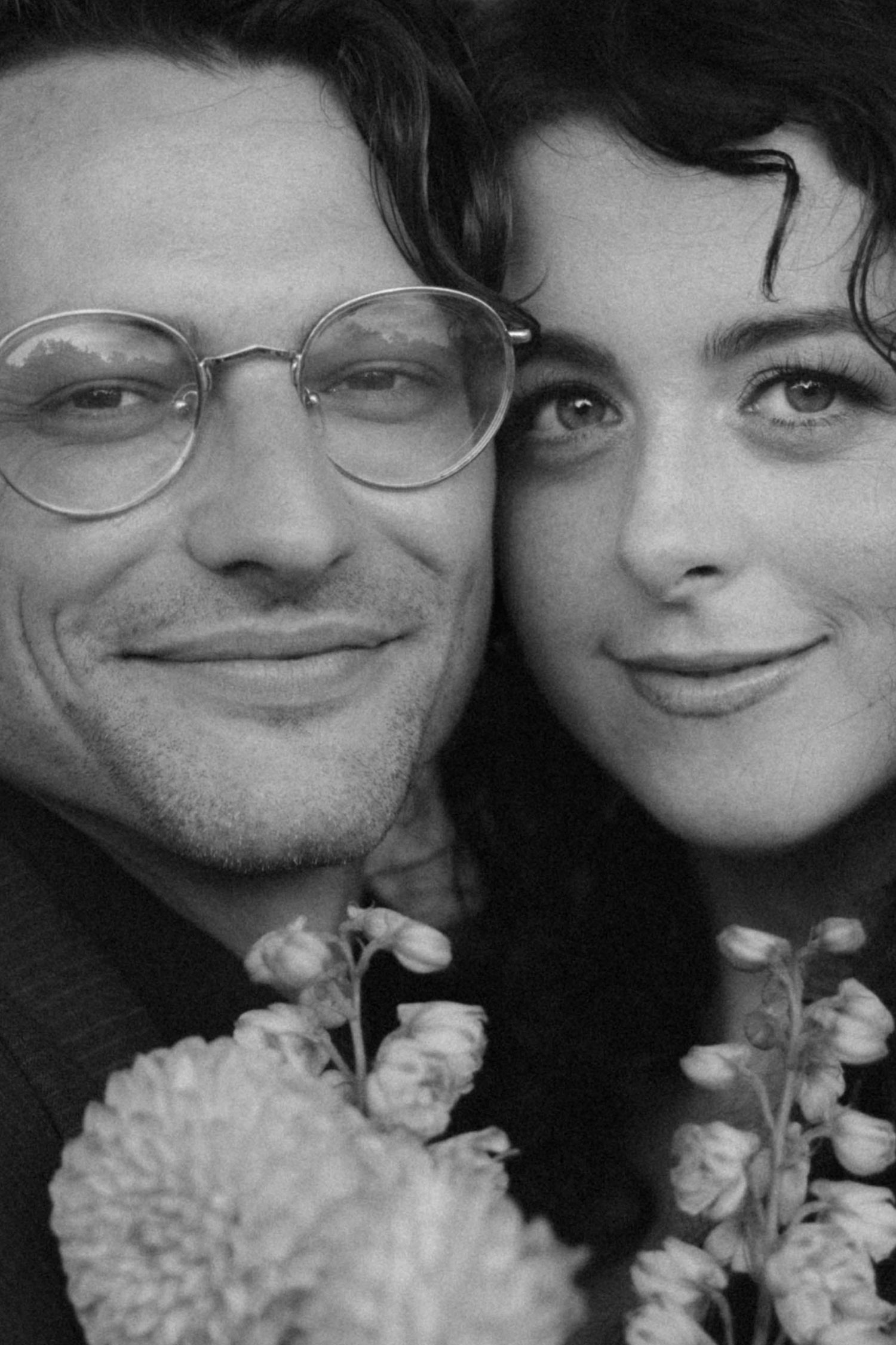 Black and white close-up portrait of a smiling couple with flowers, man wearing round glasses.