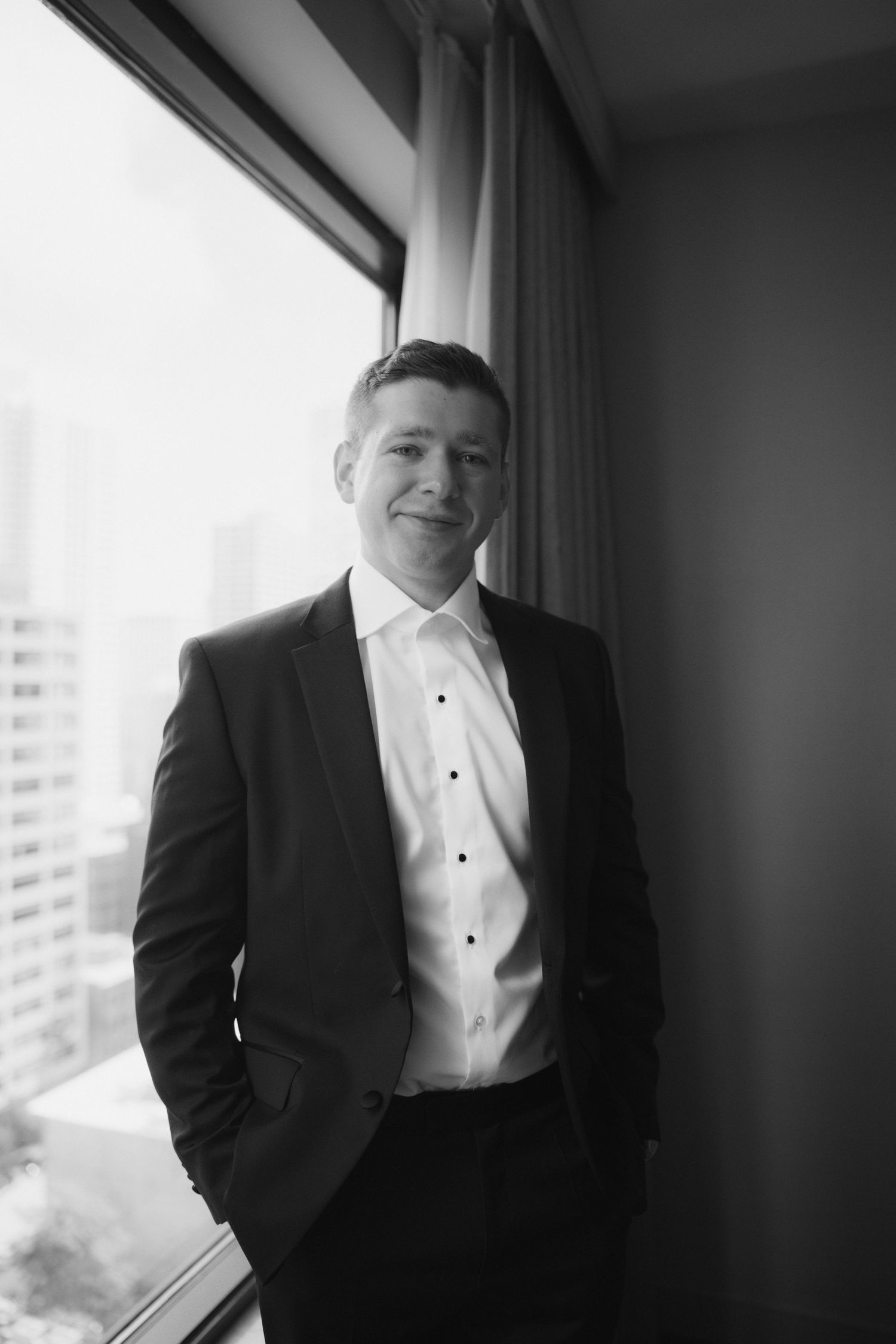 Black and white portrait of a smiling man in a suit standing by a window with city skyline views.