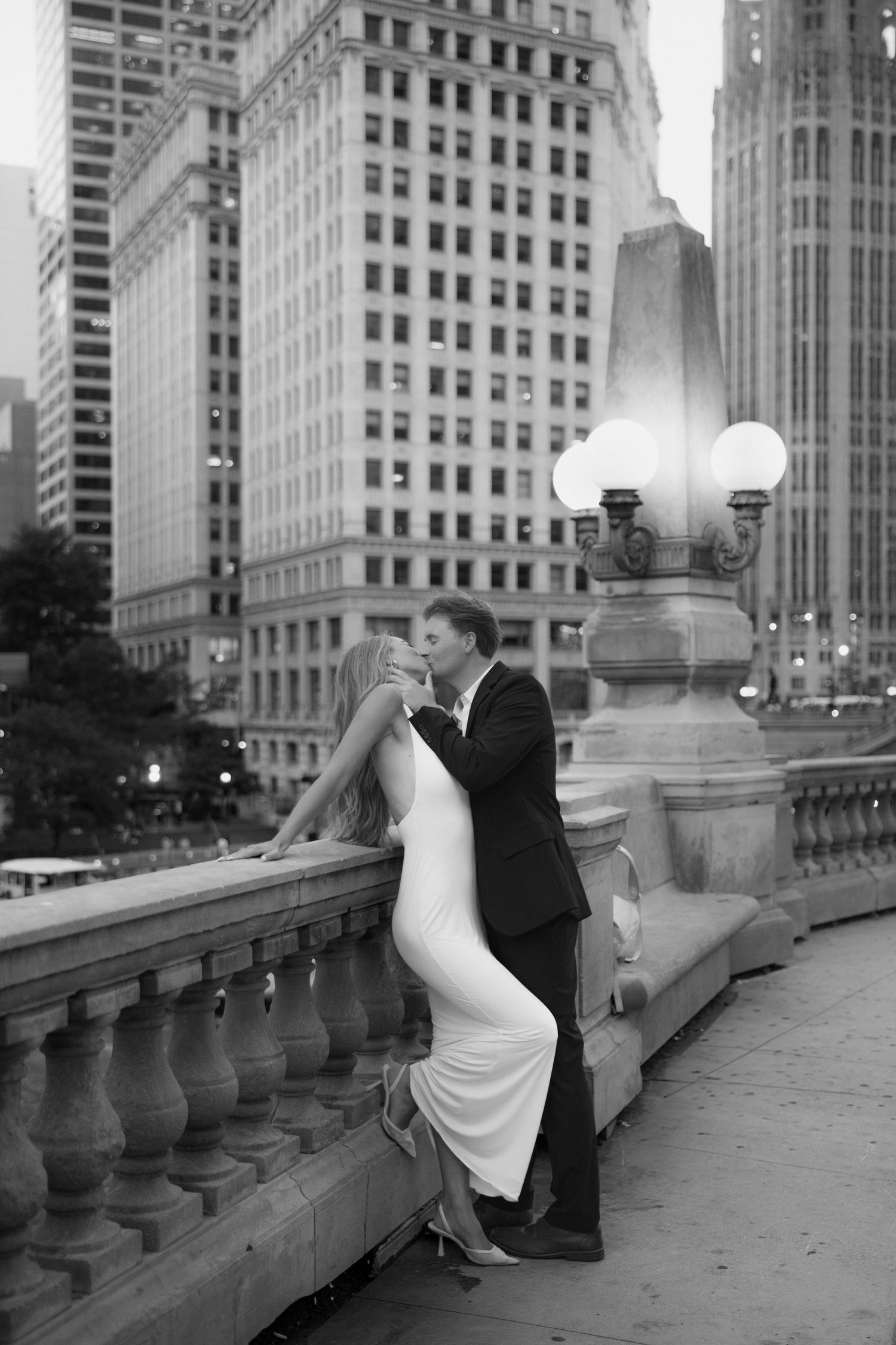 Black and white photo of couple kissing on Chicago bridge, woman in white dress, city buildings in background.