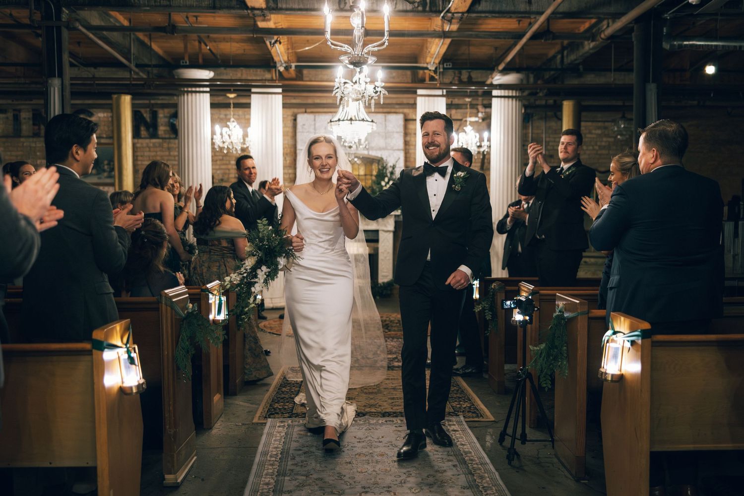 Wedding reception guests dance and celebrate on the dance floor under elegant chandeliers in an indoor venue.