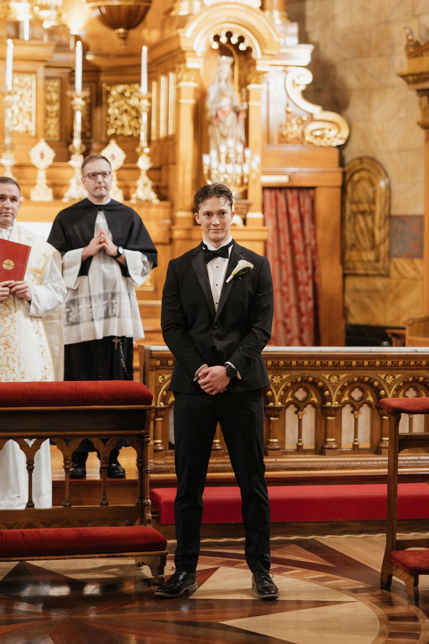 Wedding party members seated in ornate church pews with golden decor and chandeliers.