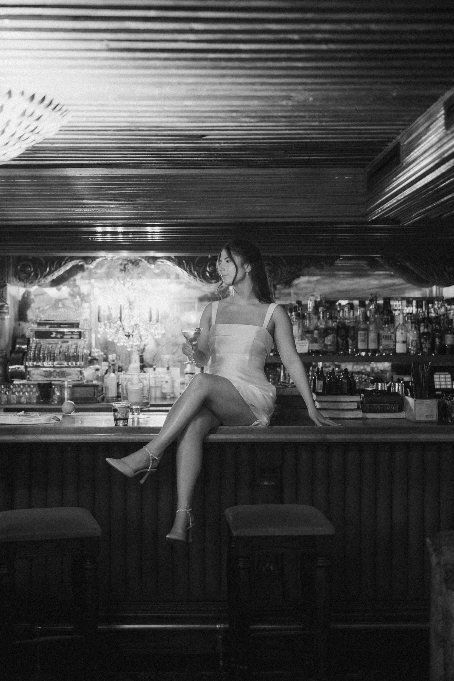Black and white panoramic photo of a dimly lit bar interior with empty stools and bottles displayed on shelves.