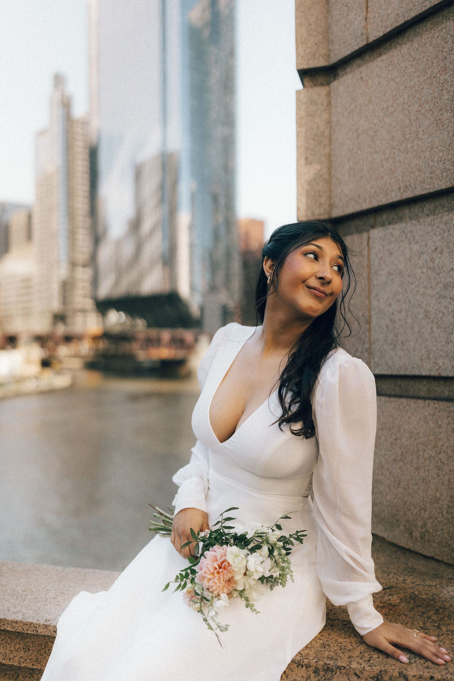 A person in a white long-sleeved wedding dress sits elegantly against a stone wall with city views.