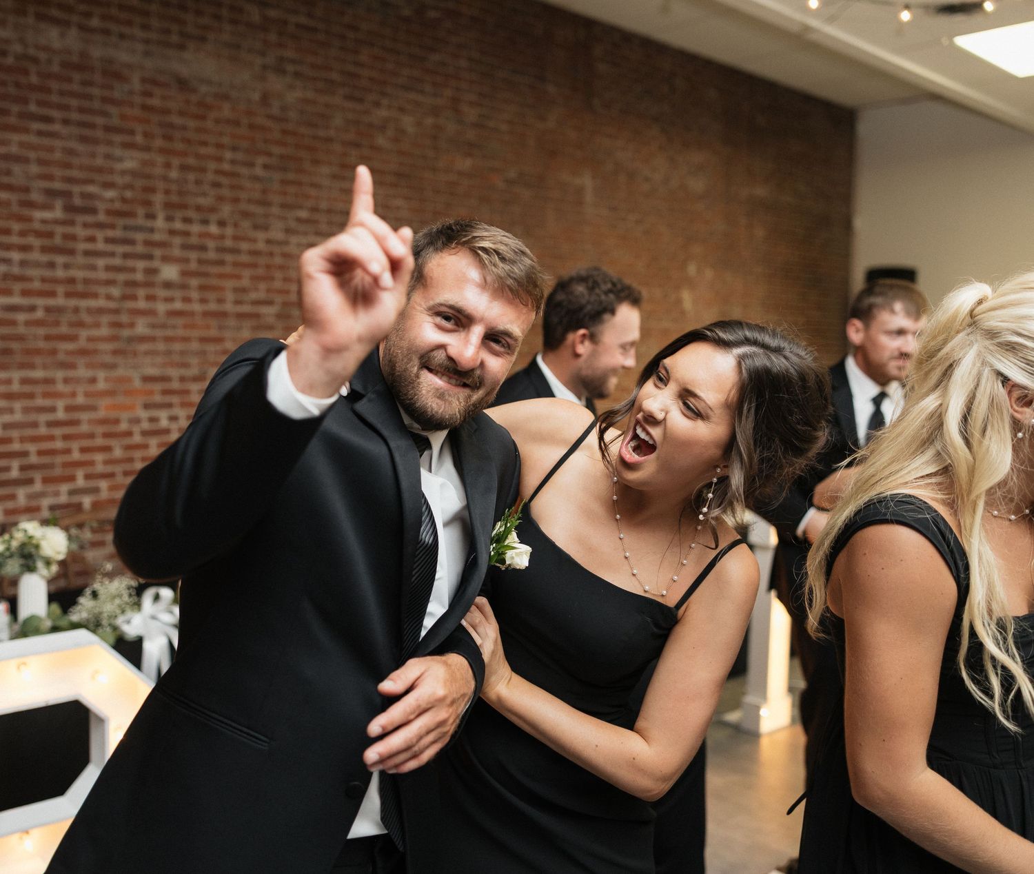 Wedding party celebrating with joyful expressions and laughter at an indoor reception with brick walls.