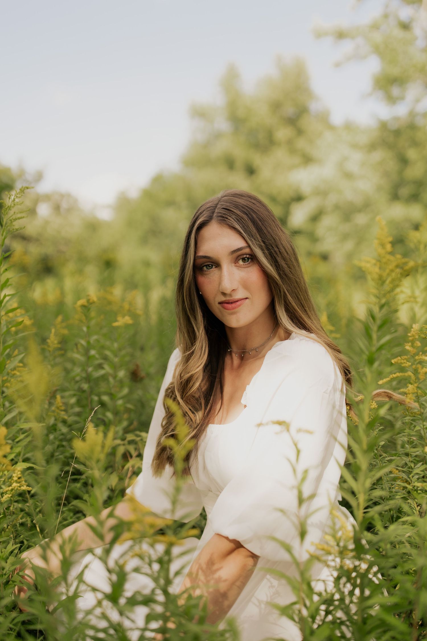 Woman in white dress poses gracefully among tall yellow wildflowers in a sunny outdoor setting.