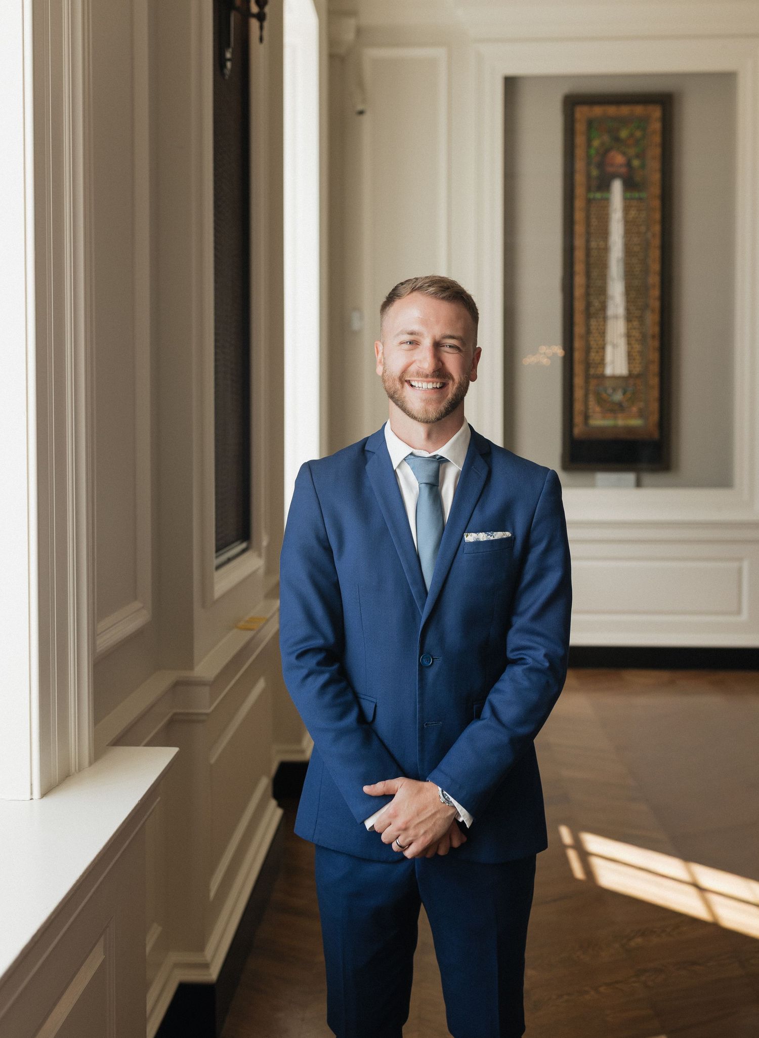 Professional in a navy blue suit smiles warmly in an elegant hallway with white walls and natural lighting.