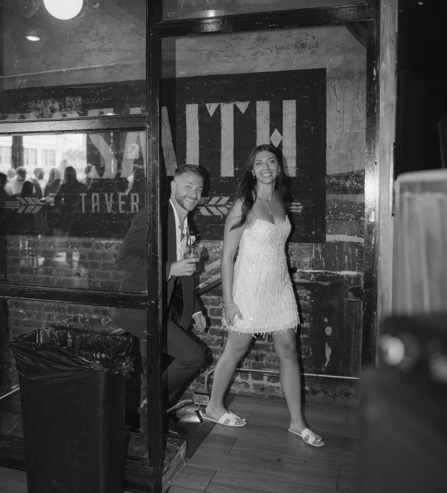 Black and white photograph showing two people posing in a casual setting near a brick wall with signage.
