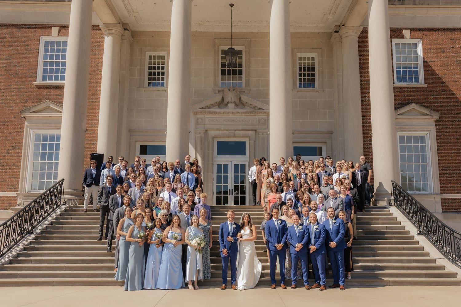 Wedding party poses on grand brick building steps with elegant columns and white trim in classic formal attire.