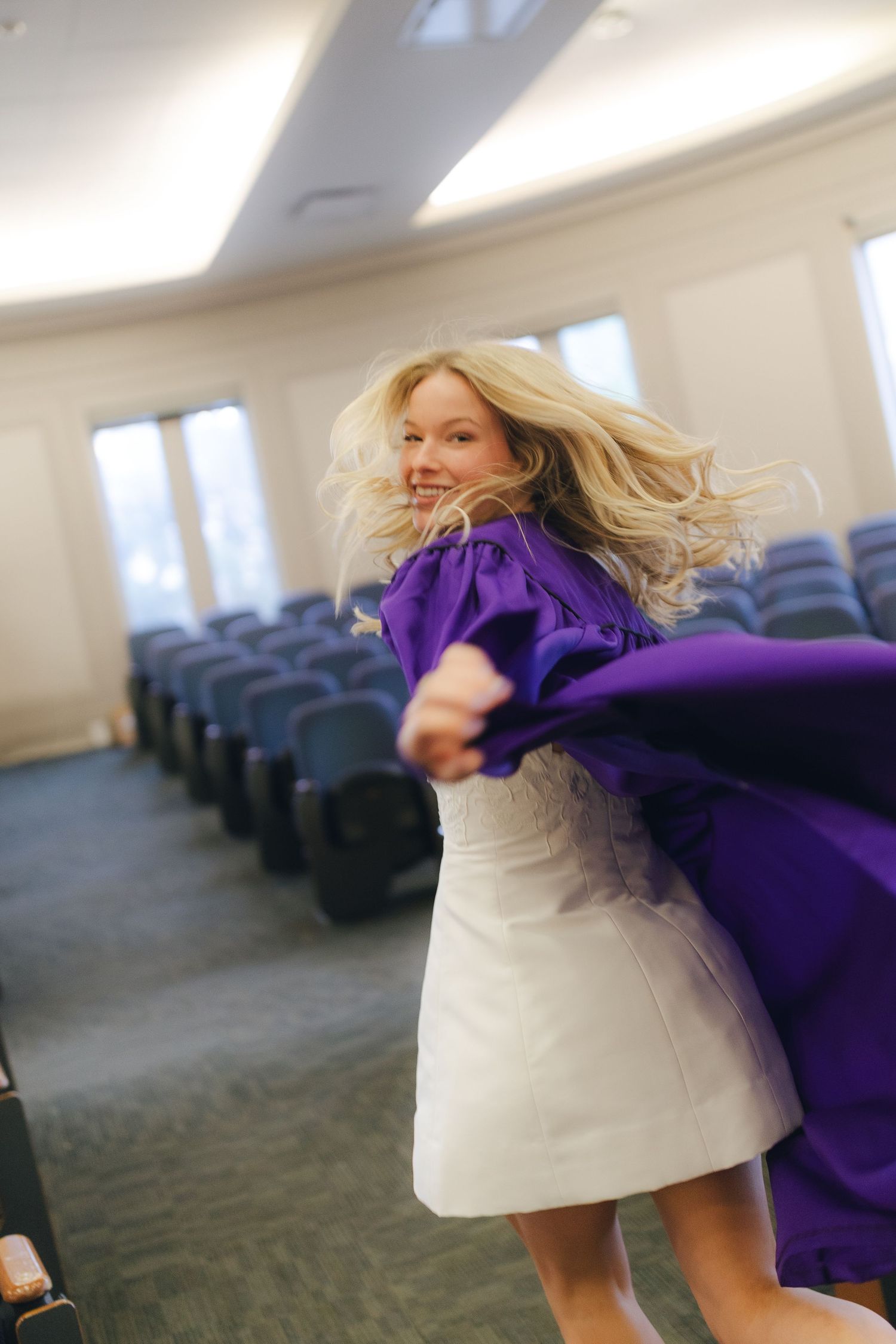 A person in a white dress spins around with a flowing purple cape in an empty conference room.