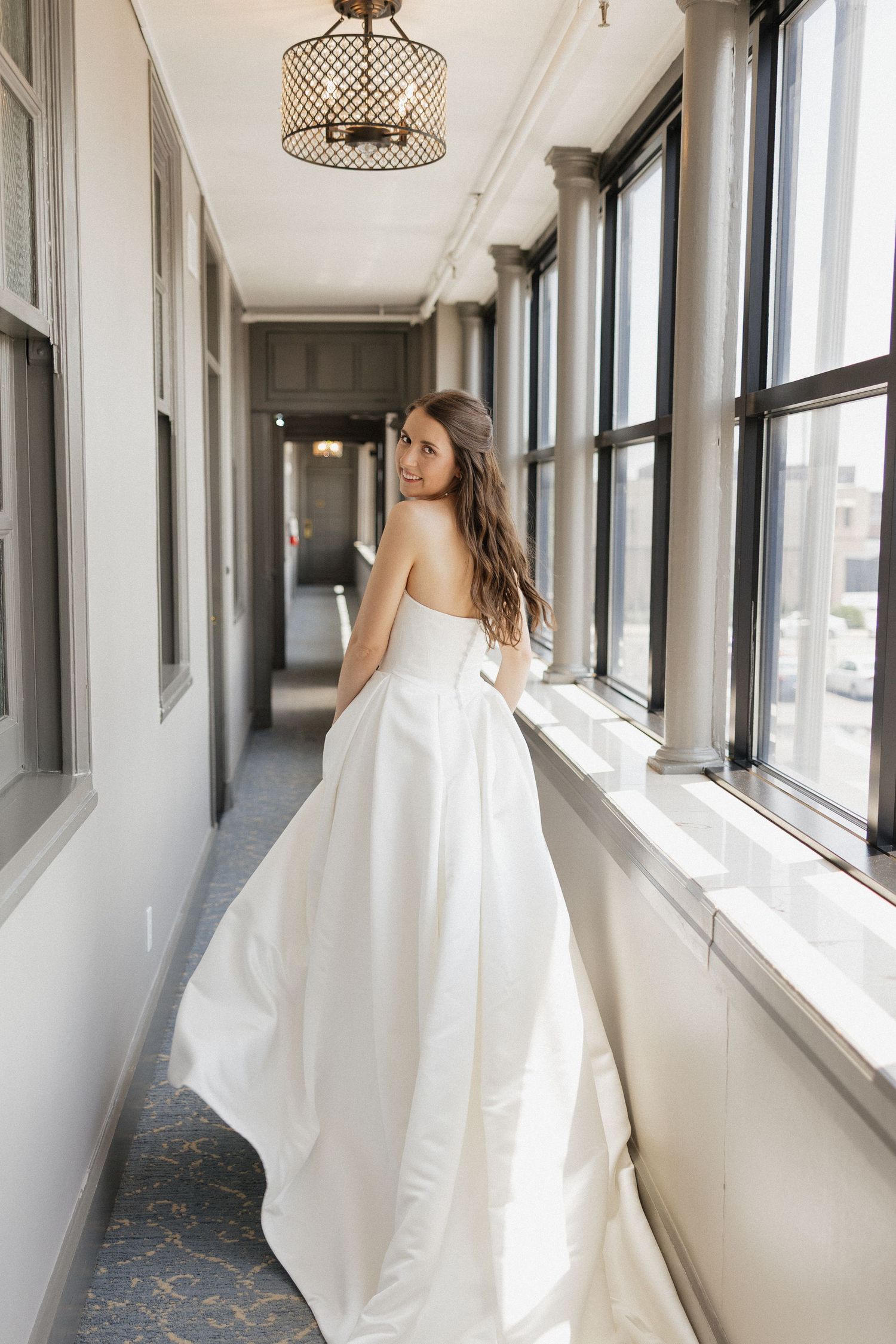 Bride in strapless white ball gown standing in elegant hallway with large windows, looking back over shoulder.