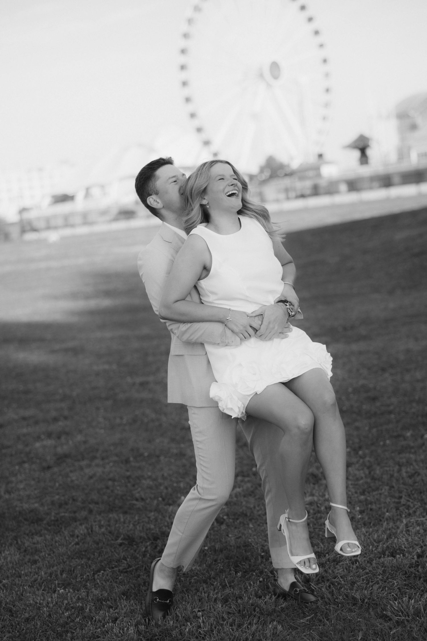 Joyful couple laughing as man lifts woman near Ferris wheel in black and white photo.