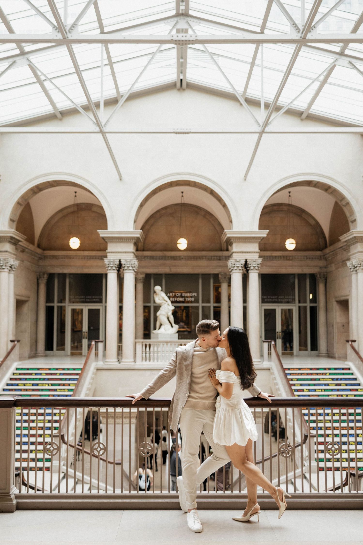 Couple sharing a romantic moment on a balcony inside a grand neoclassical museum with arched columns.