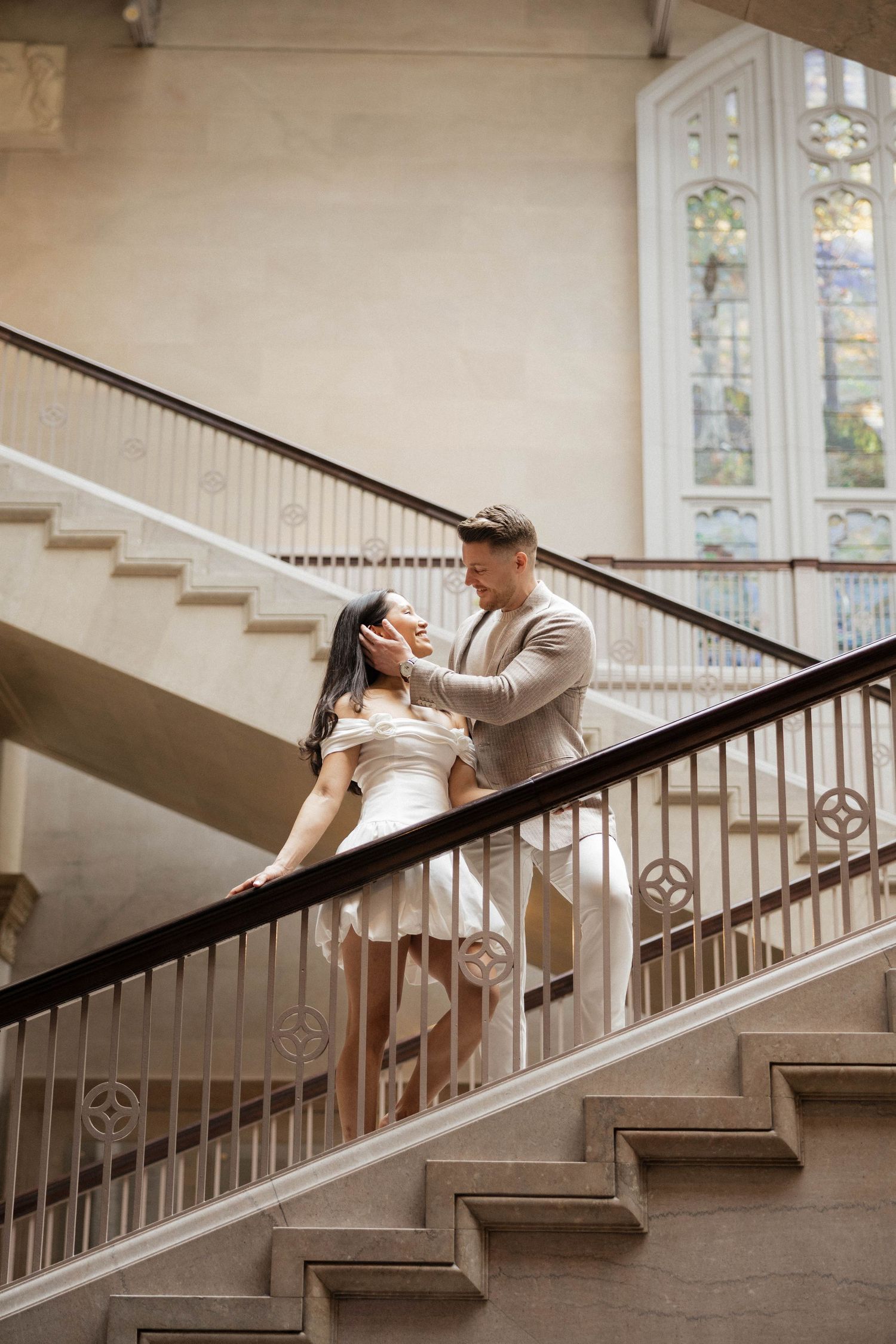 Couple sharing a tender moment on elegant staircase with ornate stained glass window backdrop.