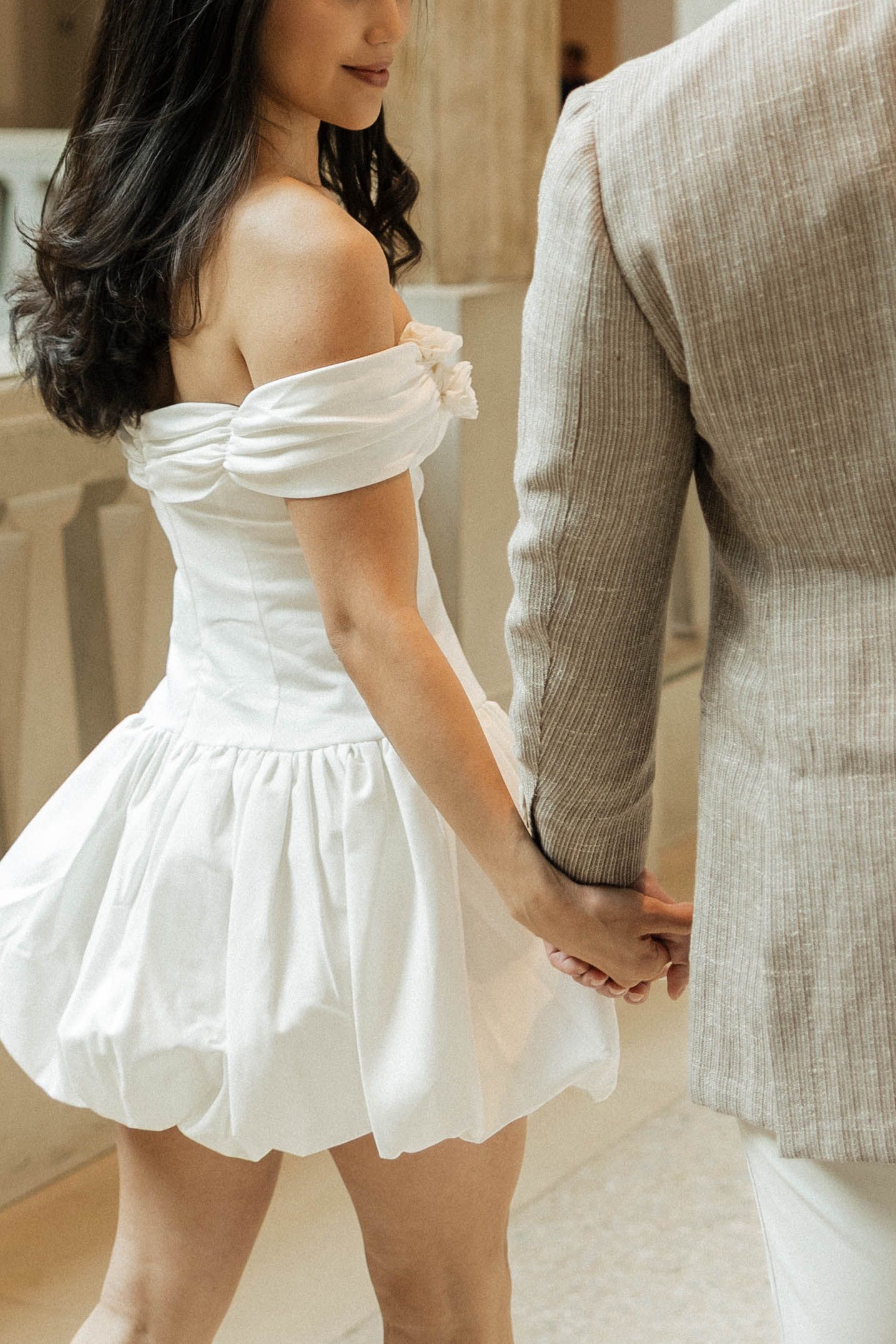 A woman in a white off-shoulder bubble mini dress holds hands with a man in a beige suit.